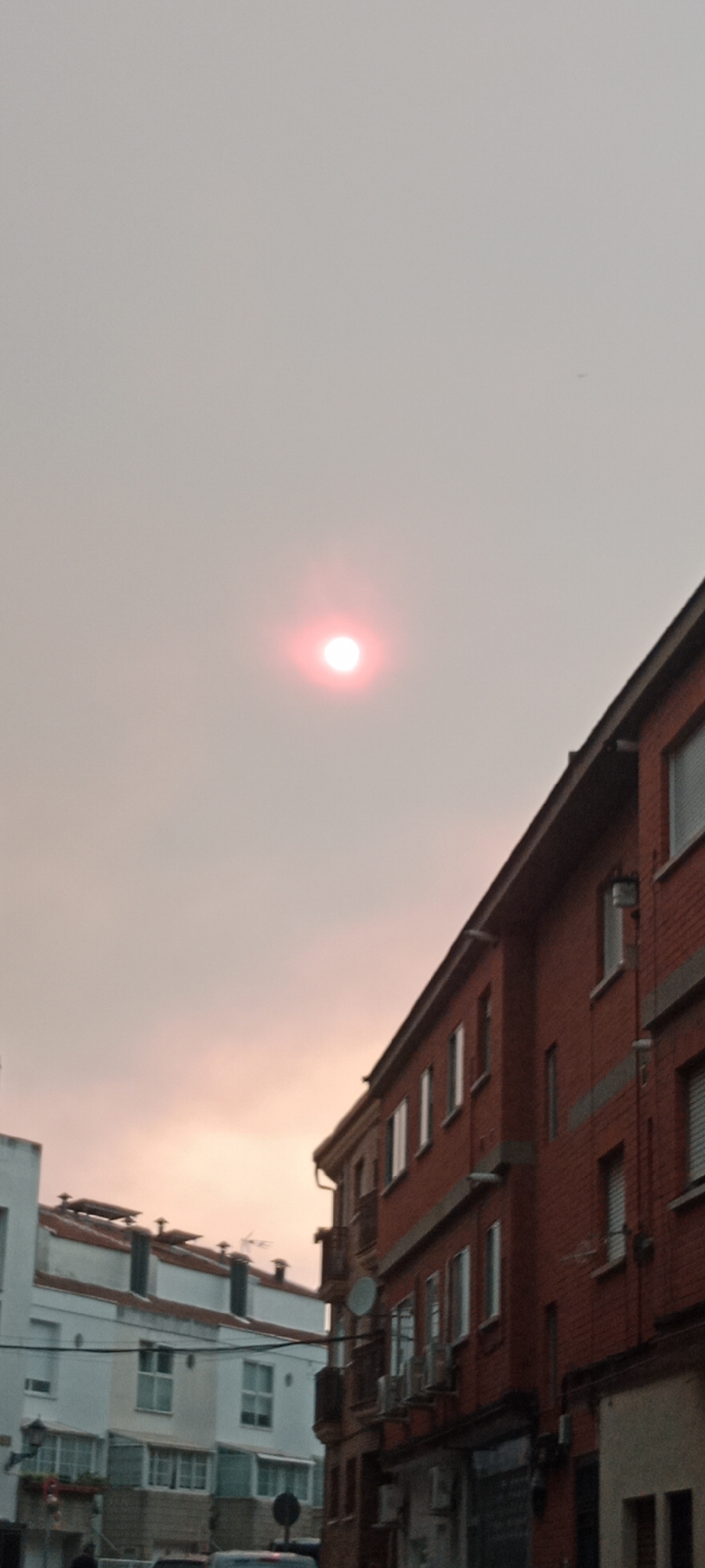 The image depicts an urban scene in Galapagar, Spain, under a hazy, pinkish sky. The sun, appearing as a bright, reddish-pink disc, is visible in the upper central portion of the frame, its light diffused by atmospheric conditions, possibly smoke or dust. In the foreground, several buildings are visible. To the right, a multi-story brick building with red bricks and white-framed windows dominates the right side of the image. Its roofline slopes downwards towards the left. Below the roof, a prominent dark gutter runs along the edge. Further to the left, a white building with a darker roofline and multiple windows is partially visible. The architecture suggests a residential or commercial area. Power lines are strung between the buildings, and air conditioning units are mounted on the exterior walls of the brick building. The overall impression is one of a slightly muted or filtered light, characteristic of a sky affected by haze or smoke. No people are clearly discernible in the frame.