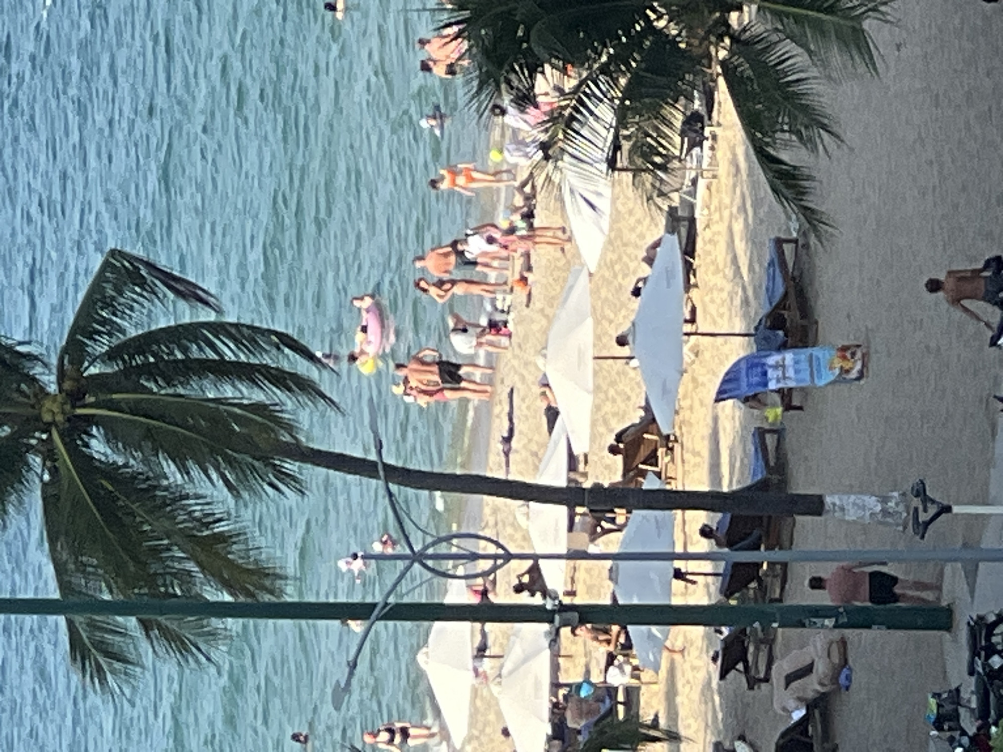 This is an eye-level, medium-range, vertical, outdoor photograph of a crowded beach scene in Nha Trang, Vietnam, taken on a sunny day. The turquoise water of the ocean is visible in the background, with gentle waves. Several people are in the water, some swimming and others floating on inflatable toys. The sandy beach is populated with numerous individuals, many of whom are sunbathing on lounge chairs or towels under white umbrellas. A few people are standing near the water's edge.
In the foreground, a tall palm tree with dark green fronds partially obscures the view on the left side of the image. A dark green pole with decorative metalwork and a lamp fixture is positioned centrally, with another similar pole to its right.
On the sand, several lounge chairs are arranged, with some occupied by people. A surf-style board with colorful graphics stands upright near the center. Further back, more people are visible, some clustered together. In the bottom left corner, the edge of a dark motorcycle is visible. The overall atmosphere appears to be one of leisure and recreation.