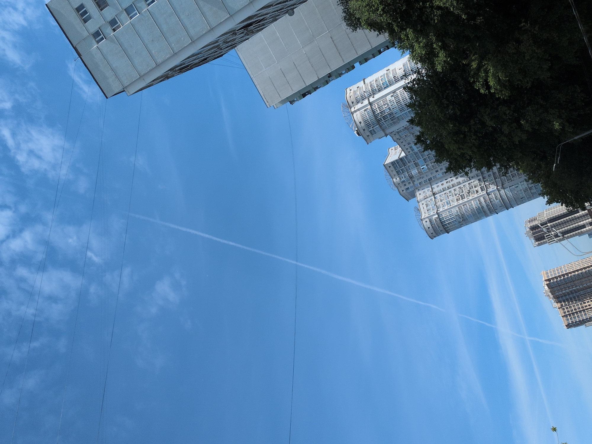 The image, taken in Moscow, Russia, presents a low-angle view directed upwards towards the sky. Modern, high-rise buildings are visible along the periphery of the frame. Green foliage is present in the upper right quadrant, suggesting a nearby tree. Several thin wires or cables extend from the top left corner downwards into the blue sky. A contrail bisects the sky horizontally. The image suggests an urban environment with a clear, sunny day.
