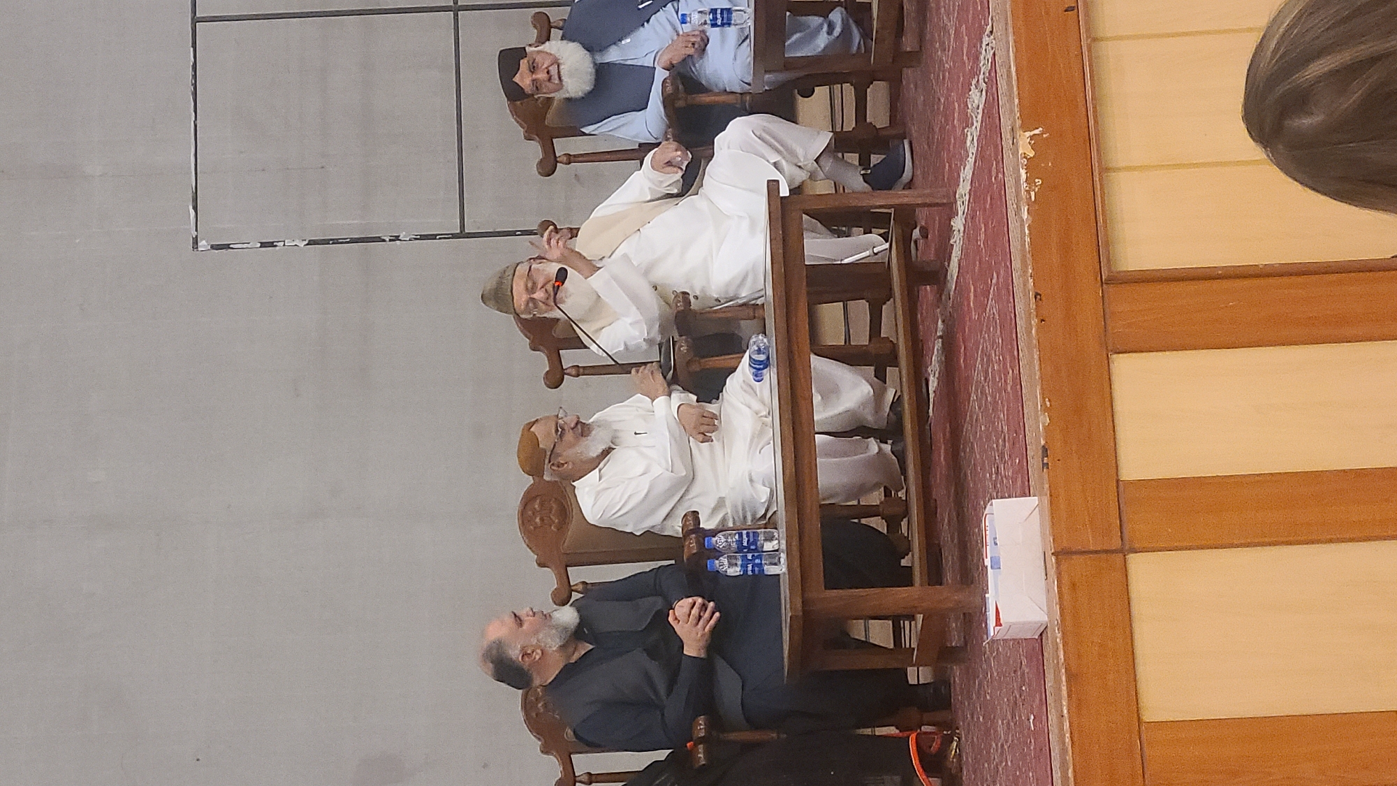 A vertically oriented, medium shot reveals a group of men seated in wooden chairs, engaged in a discussion or presentation. The location appears to be within a building, potentially in the city of Quetta City Tehsil, Pakistan.
The men are dressed in a mix of traditional and formal attire. Several of them wear white robes, while one man is dressed in a dark suit. Beards are visible on most of the men. Some of them hold water bottles on the tables in front of them.
The background consists of a wall, and a reddish carpet or rug on the floor. A woman's head is visible in the corner.