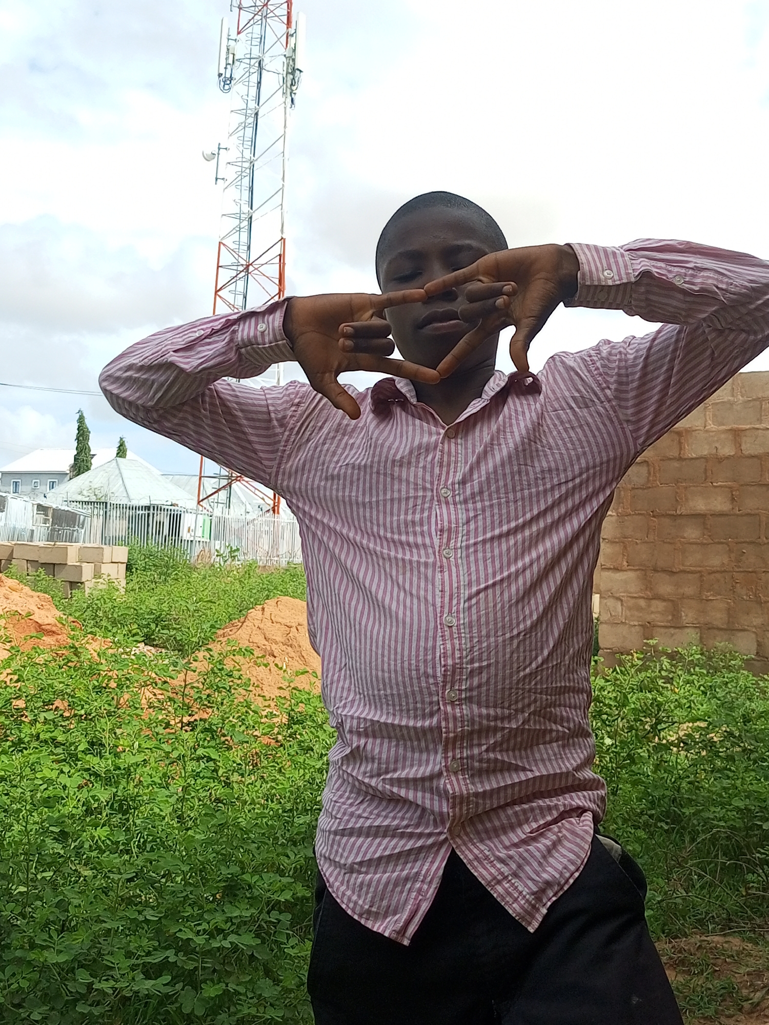 A young person, with dark skin and short hair, stands in front of a brick wall and green foliage. They are wearing a pink and white striped button-down shirt and dark pants. Their hands are held up to their face, forming a heart shape with their fingers. In the background, a tall telecommunications tower is visible against a cloudy sky. There are also some buildings, a fence, and stacks of bricks. The ground appears to be a mix of sand and vegetation. The overall scene suggests an outdoor location in Tempure, Nigeria.