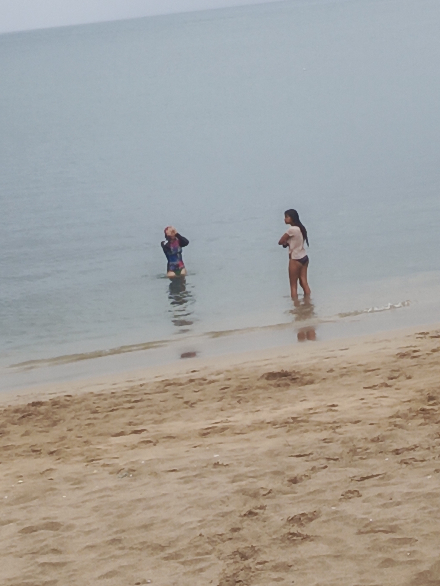 Two individuals are wading in the shallow waters of a beach. One person, wearing a full-body swimsuit with a colorful pattern, is positioned further back in the water, appearing to be adjusting their goggles or sunglasses. The second person, closer to the shore, is wearing a white t-shirt and dark bottoms, with their arms crossed. The environment is a beach with sandy terrain and calm, overcast water. The scene suggests a casual day at the beach in Juan Griego, Venezuela.