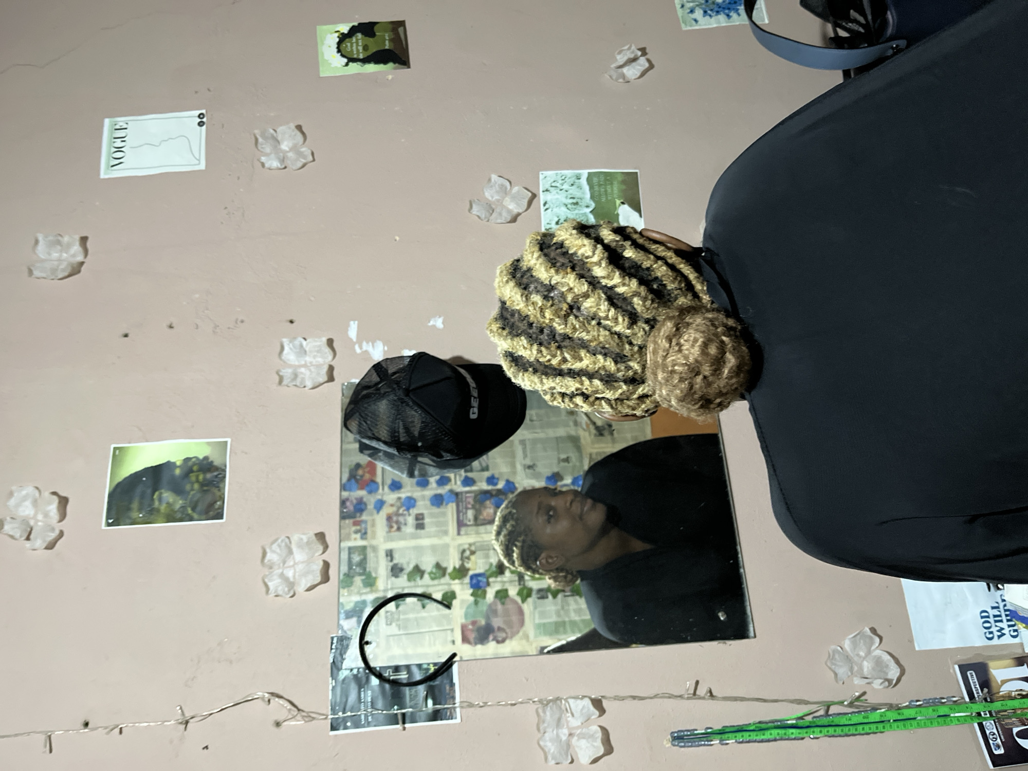 The image depicts an indoor scene, likely a bedroom or personal space, with a person facing away from the camera, looking into a mirror. The walls are a pale pink hue and are decorated with several small, white, flower-shaped ornaments, and various posters and images. Visible posters include a "VOGUE" magazine cover, a picture of a bird, and motivational quotes. In the mirror, a woman with braided hair is visible, wearing a black top. A black baseball cap with "GEEZ" printed on it is hanging on the wall above the mirror. To the left of the mirror, a string of fairy lights is visible. In the foreground, the person facing away from the camera has braided hair styled into a bun. Books and papers are visible on a surface below the mirror. The overall impression is that of a personal, somewhat cluttered, decorative space. The location is identified as Rafin Par, Nigeria.
