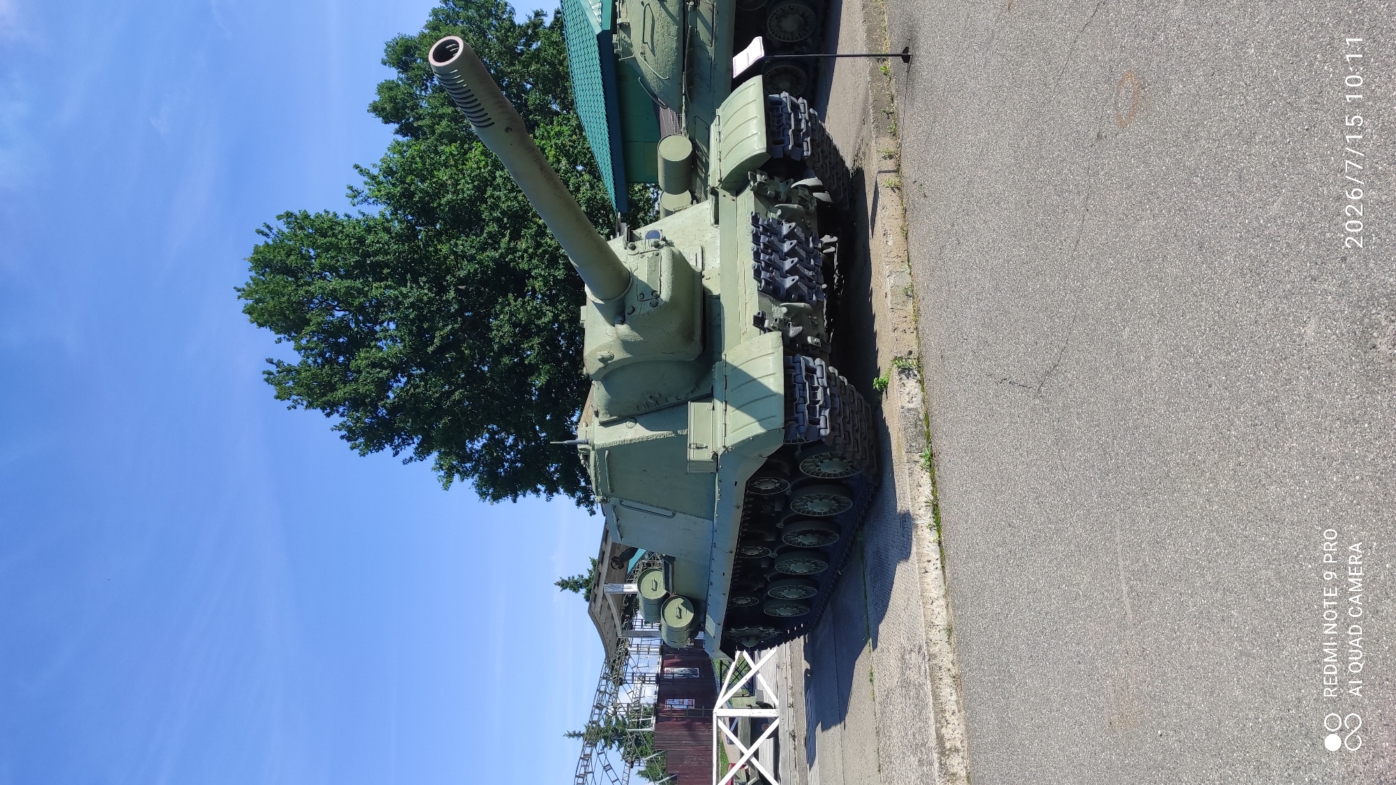 A green self-propelled artillery gun, likely a Soviet-era 2S3 Akatsiya, is on display outdoors. It is positioned on a concrete platform next to an asphalt road. The gun is oriented towards the right side of the frame, with its long barrel prominently featured. In the background, there are trees and a green-roofed building. A white fence is visible to the left, and other military vehicles are partially seen in the background. The sky is clear blue. The image appears to be taken during daylight hours. The bottom left of the image contains a watermark with "REDMI NOTE 9 PRO AI QUAD CAMERA". The bottom right contains a date and time stamp: "2026/7/15 10:11". This is likely a museum or a historical display.