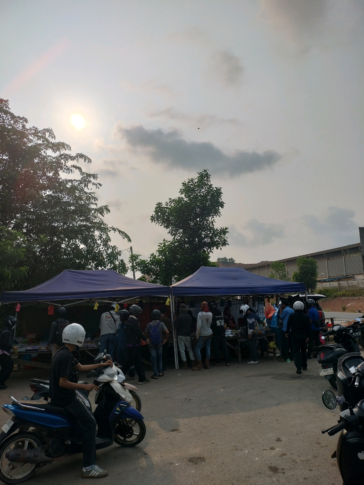 In Batam, Indonesia, a street scene unfolds with several vendors under blue tents, their stalls laden with goods. A group of people, likely customers and vendors, are gathered around the stalls. On the right side of the frame, motorcycles are parked, some with riders or passengers. The sun is low in the sky, casting a warm glow and creating lens flare. Trees and a building are visible in the background. The ground is paved with asphalt.