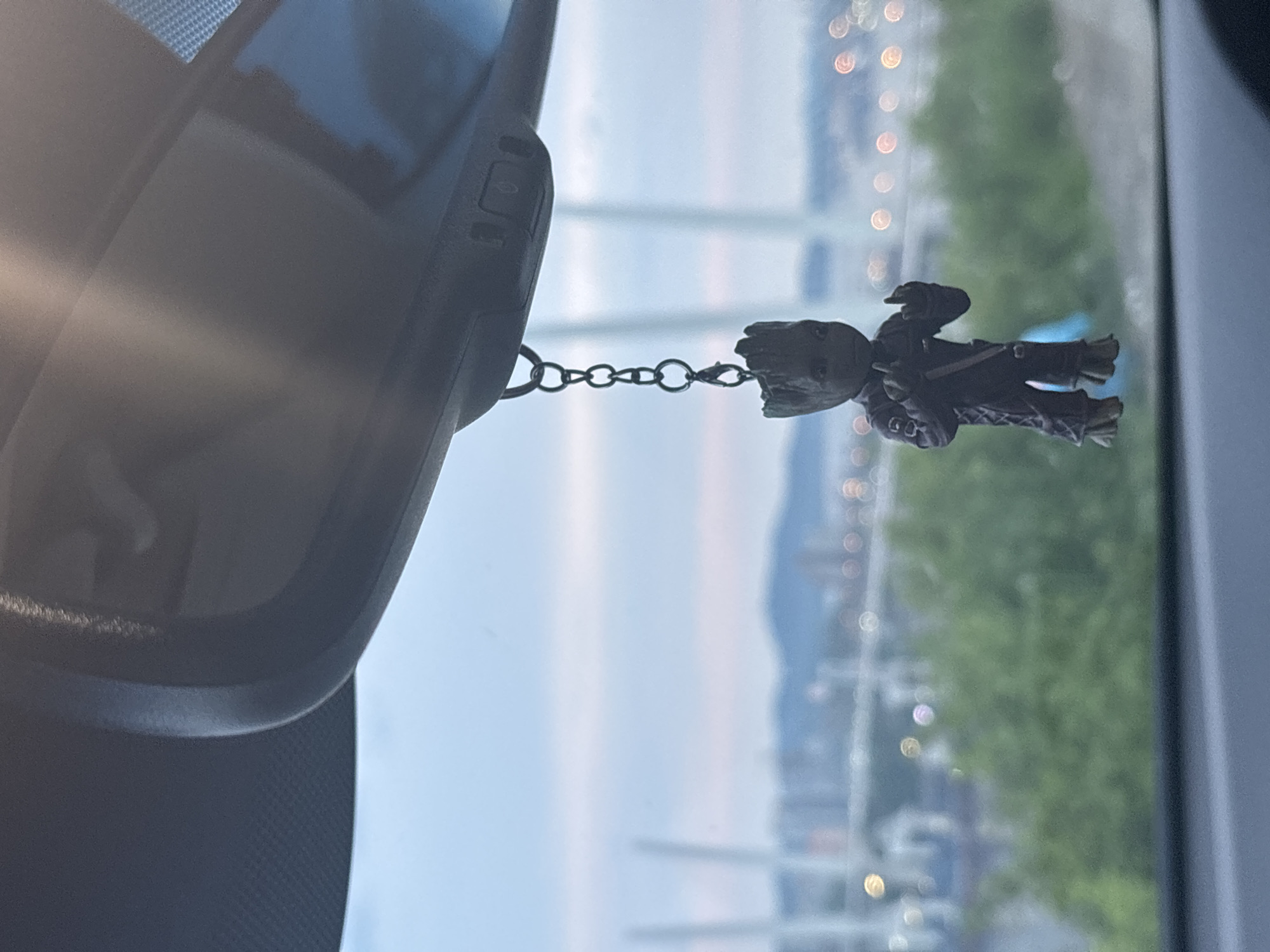 A dark, miniature figurine of Groot, a character from the Marvel Cinematic Universe, hangs from a chain from the rearview mirror of a vehicle. The figurine is detailed, with visible textures on its bark-like skin and clothing. It is positioned in the foreground, with a blurred cityscape visible in the background. The cityscape includes bridges, buildings, and green foliage, suggesting an urban environment. The sky is overcast with soft, pastel hues, indicating either dawn or dusk. The scene is likely captured from inside the vehicle, looking out through the windshield.
