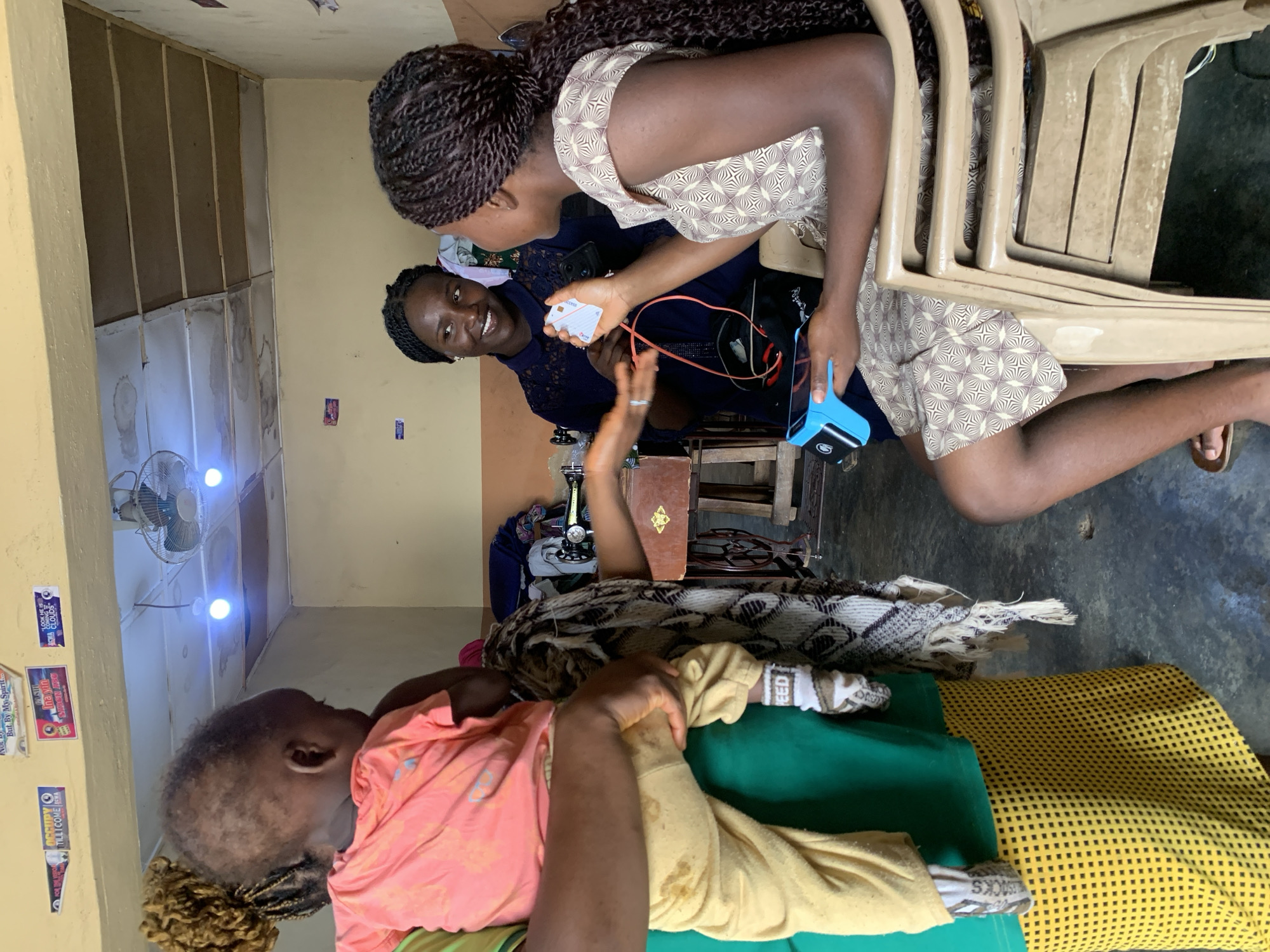 In an indoor setting resembling a small shop or workshop, three individuals are present. A woman, seen from the side and partially obscured, holds a baby. The baby, wearing a pink shirt and yellow pants, faces away from the camera. To the right, another woman is seated, holding a credit card reader in her left hand and a credit card in her right hand. She is looking and smiling at a third woman, who is standing and appears to be in conversation with the seated woman. Behind them, a sewing machine and various fabrics are visible, suggesting the location might be a tailor's shop. The walls are painted a pale yellow, and the ceiling has exposed wooden beams with some water stains. A ceiling fan is mounted on the ceiling. The floor is a concrete surface. The overall impression is a casual interaction within a functional workspace.