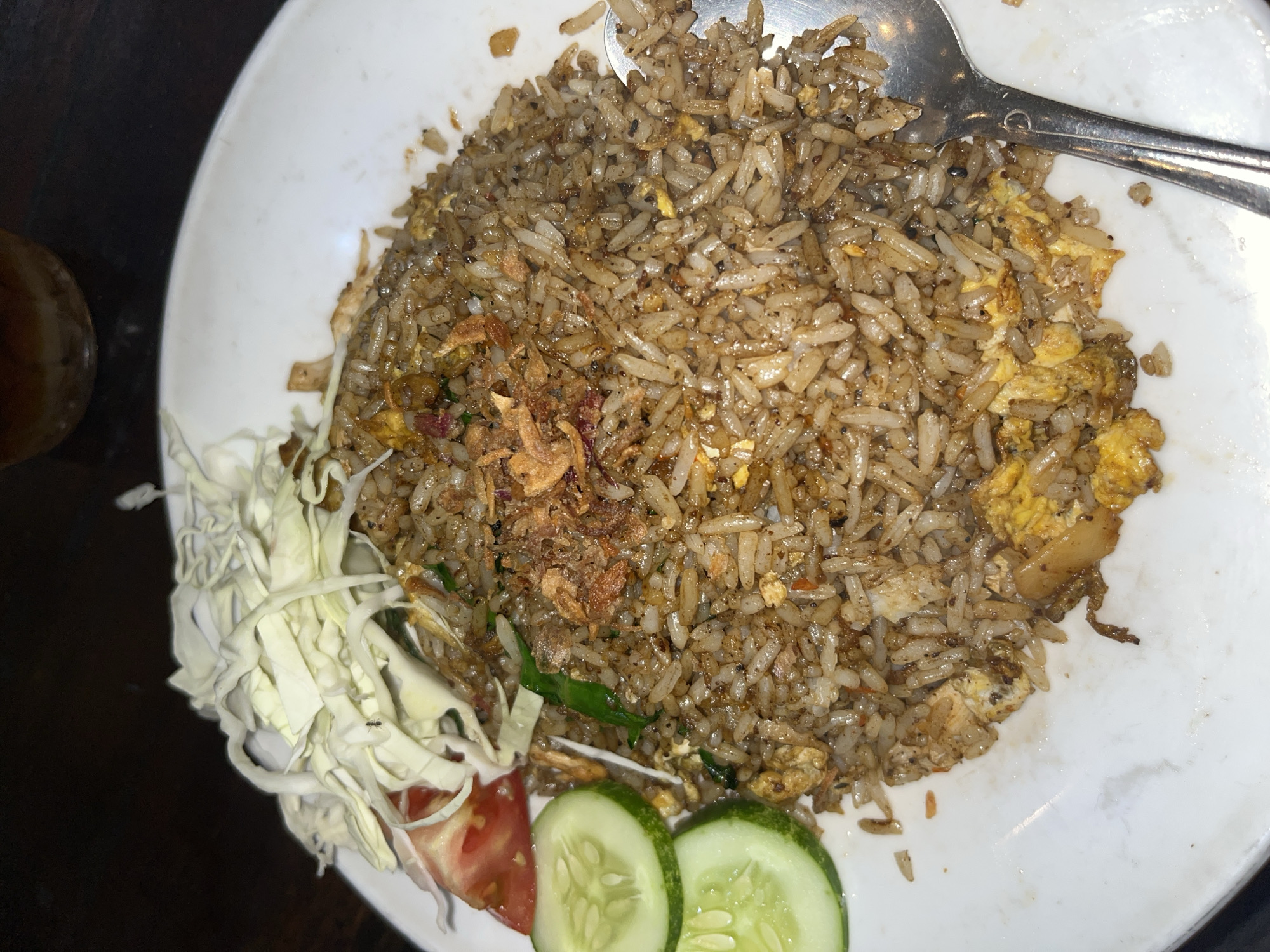 An overhead shot captures a plate of Nasi Goreng, a popular Indonesian fried rice dish, served on a white ceramic plate. The dish is a generous portion of rice, stir-fried with what appears to be egg and dark soy sauce, giving it a rich brown color. Garnishes include shredded cabbage, sliced tomato, and cucumber on the side. Crispy fried onions are sprinkled on top of the rice. A metal spoon rests on the right side of the plate, partially submerged in the rice. The background is dark, suggesting an indoor setting, possibly a restaurant in Tampingan, Indonesia, given the context.