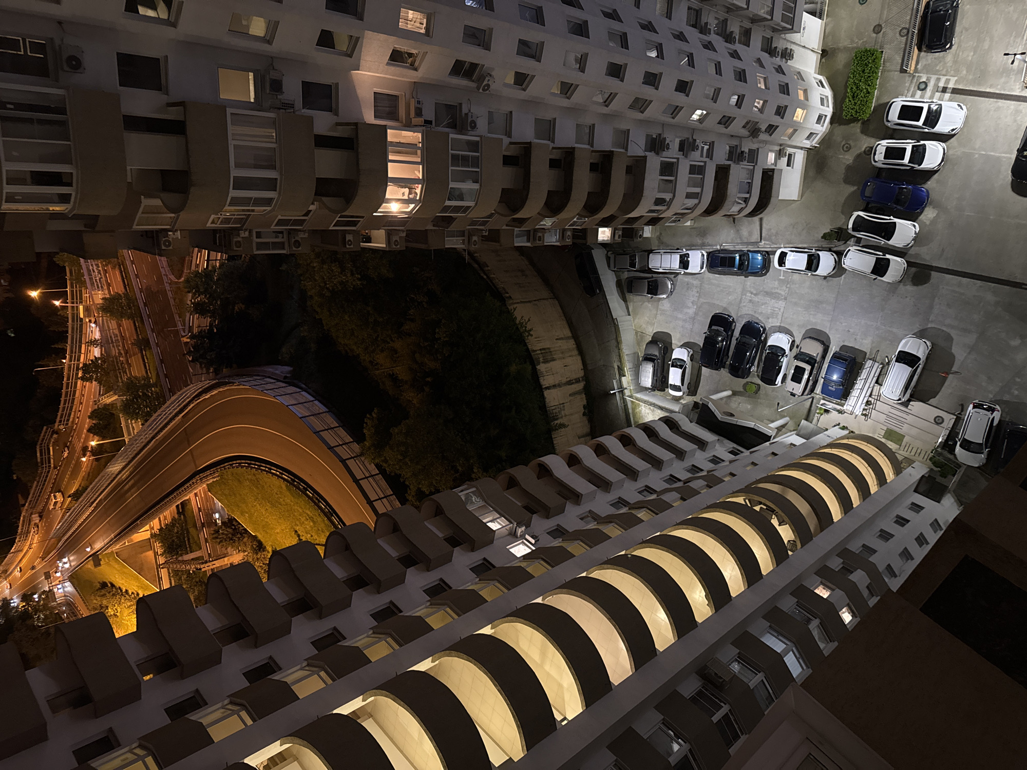 Viewed from a high angle at night in Sochi, Russia, the image showcases the interplay of architecture and infrastructure. Two tall apartment buildings dominate the frame, one on the left and the other on the right. The buildings have protruding balconies with arches. The buildings are illuminated, casting light on the balconies and the surrounding area.
Between the two buildings, a multilane highway curves. Streetlights line the highway, illuminating the roadway. Adjacent to the highway, there is a dark green grassy area.
A parking lot sits to the side of the building on the right. Several cars of various colors are parked. A small area of foliage is visible.