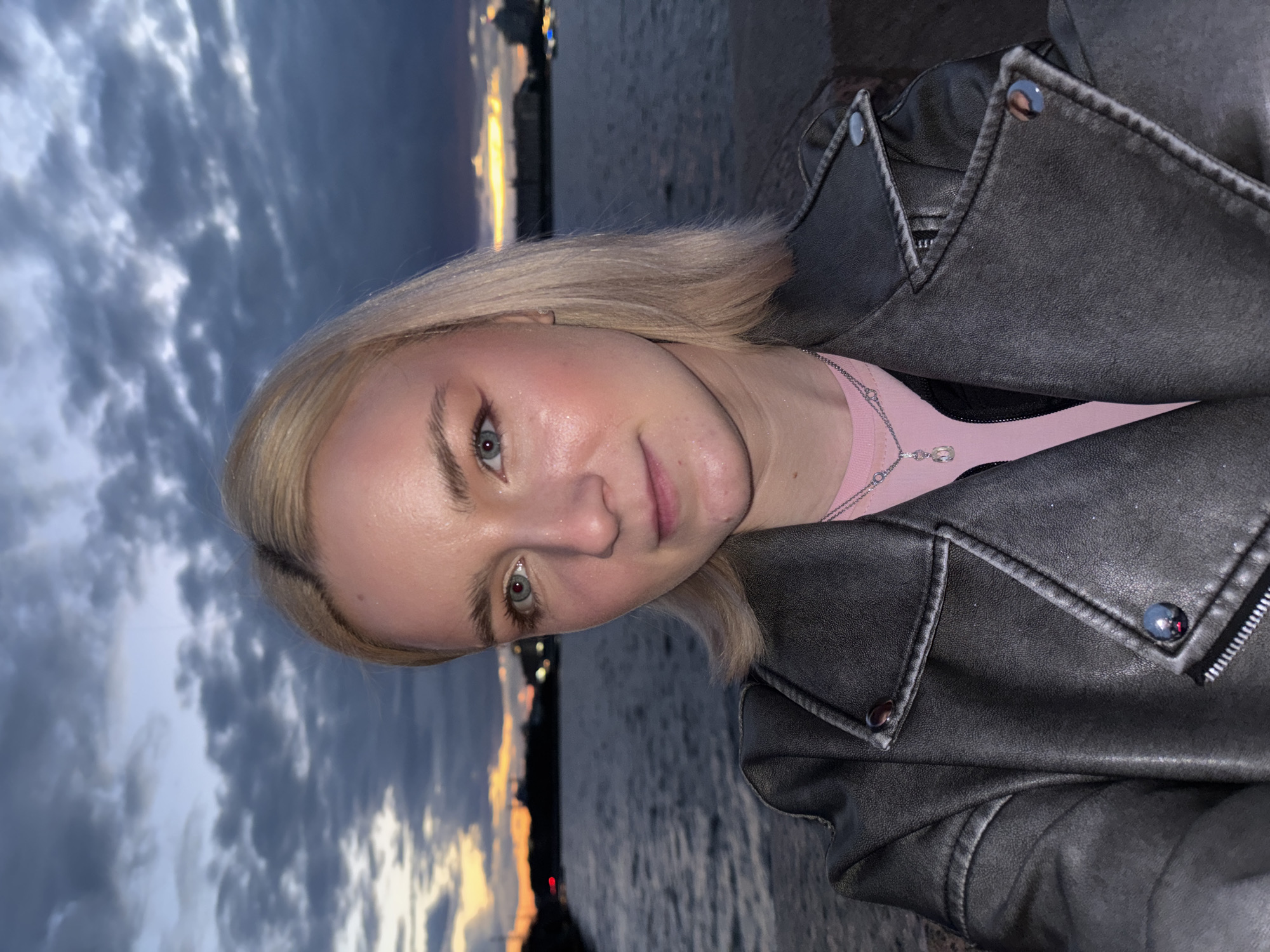 A young woman with blonde hair and light-colored eyes is pictured from the chest up, facing the camera. She is wearing a grey leather jacket over a pink shirt and a silver necklace with a crystal pendant. The background depicts a body of water, likely a river or canal, with a city skyline visible in the distance during twilight. The sky is filled with dramatic clouds and the warm glow of the setting sun. The lighting suggests it is dusk in Saint Petersburg, Russia, possibly along the Neva River or one of its canals, given the presence of water and urban infrastructure.