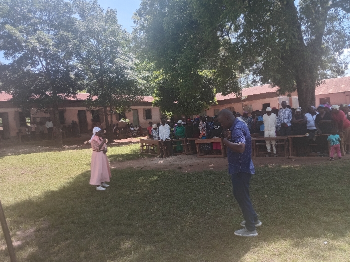 The image depicts an outdoor gathering in the Idakho South ward, Kenya, under a clear sky. In the foreground, a manicured grassy area is visible. In the midground, a crowd of approximately 30-50 individuals, primarily adults, is assembled. Some are standing, while others are seated on wooden benches arranged in rows.
On the left side of the midground, a woman wearing a light pink long dress, a white head covering, and white shoes stands, facing slightly right. She holds a small, indistinct object in her hands, positioned near her chest. On the right side of the midground, a man wearing a blue short-sleeved collared shirt with white polka dots, dark trousers, and grey sneakers stands with his head slightly bowed, his hands clasped together, holding a small dark object.
Among the gathered crowd, a man in a white tunic and white head covering is prominent on the right, and a young child in a light blue top and pink trousers is held by an adult on the far right. The crowd generally faces the left/center of the frame. In the background, multiple single-story buildings with light-colored walls and reddish-brown roofs are visible, partially obscured by large, deciduous trees. The scene suggests a community event or address.