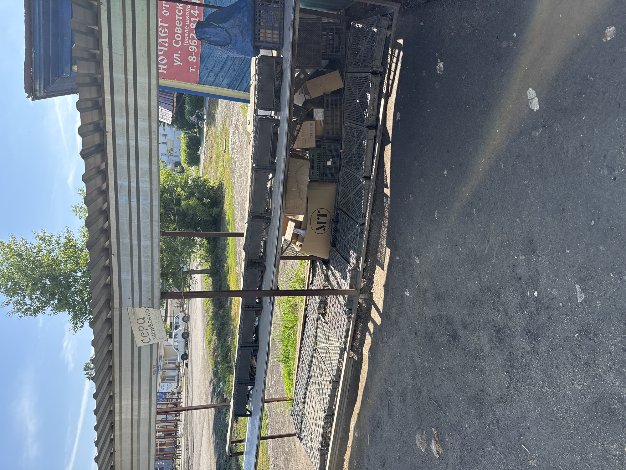 An outdoor market stall with a corrugated metal roof stands in the sun. The stall has two shelves filled with dark grey plastic crates and cardboard boxes. One sign on the stall reads "сера лиственнчная". Another sign, partially visible, advertises accommodation with "НОЧЛЕГ ОТ..." and a phone number. In the background, two white cars are parked near a building, and a person rides a bicycle on a paved path. The ground in front of the stall is dark asphalt. The scene appears to be in the town of Soloneshnoye, Russia.