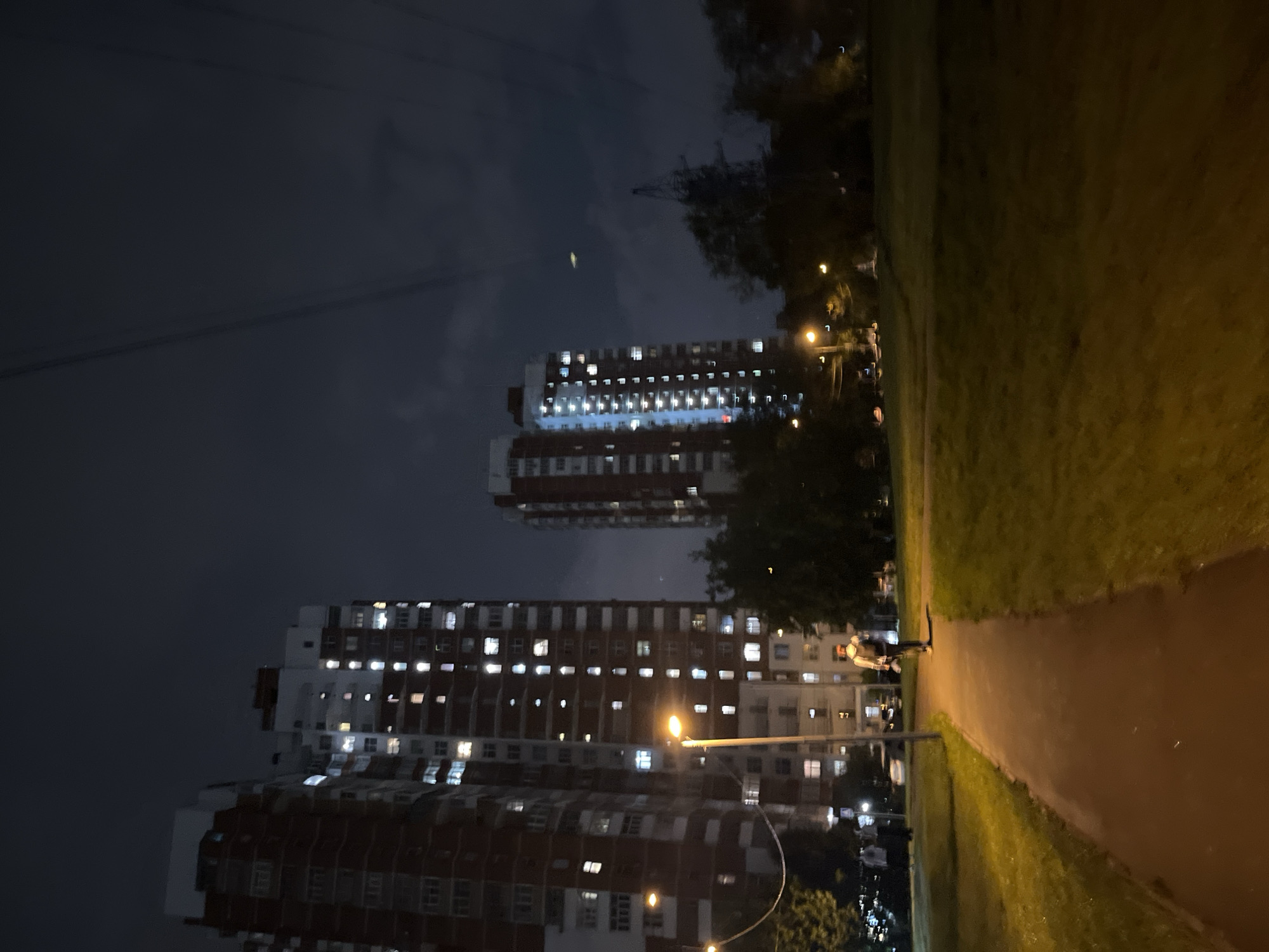A nighttime scene in a residential area of Moscow, Russia, features several tall apartment buildings with illuminated windows. In the foreground, a paved walkway curves through a grassy park area. A single person, with a backpack, walks away from the camera on the walkway. Streetlights cast a warm glow on the path and surrounding vegetation. The sky is dark and cloudy, with power lines visible. The architecture of the buildings suggests they are Soviet-era panel buildings, typical of many Moscow neighborhoods.