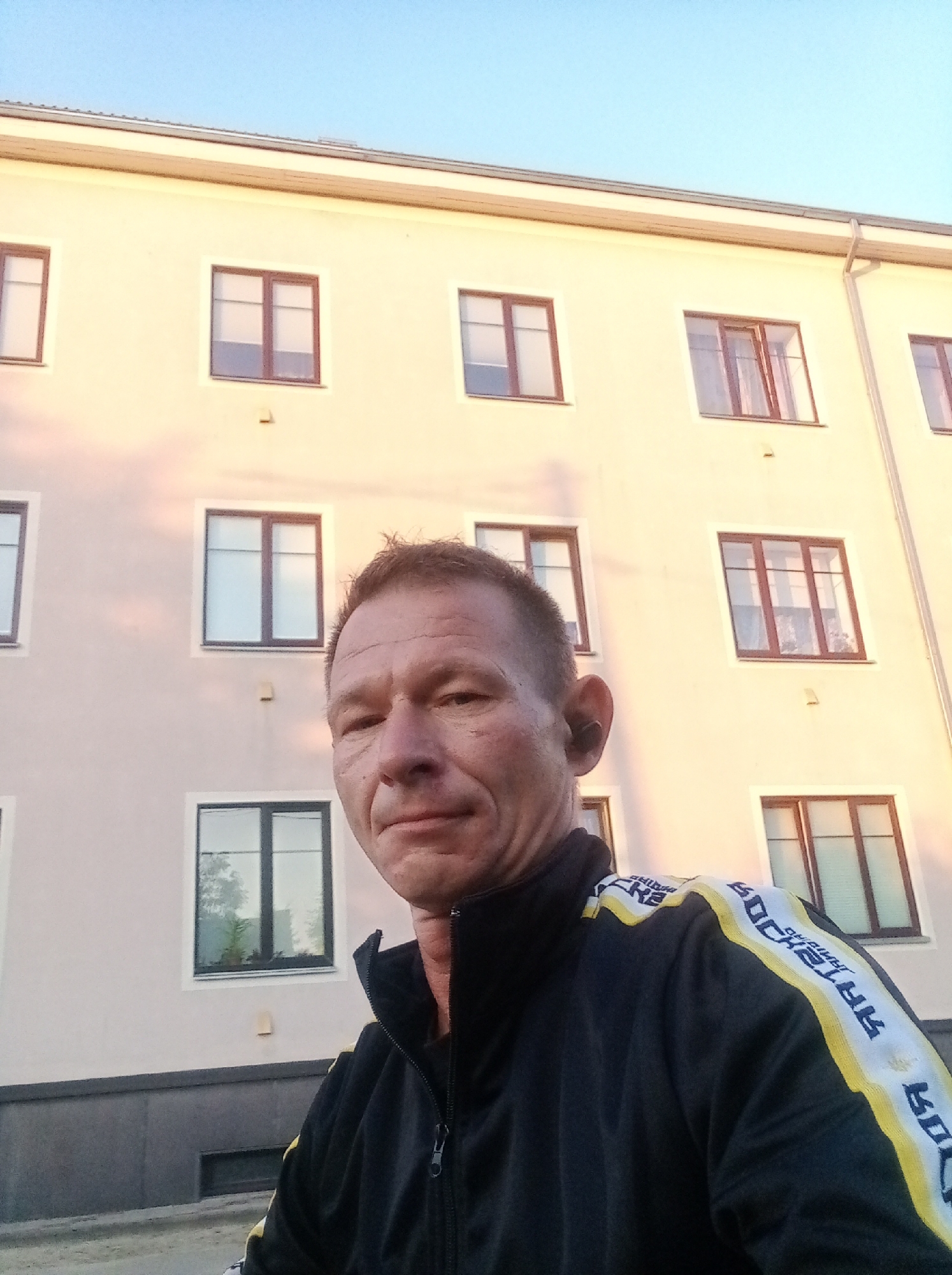 A low-angle, wide shot captures a man in a black tracksuit with yellow and white stripes on the sleeves, standing in front of a light-colored building with multiple windows. The man, positioned in the foreground and to the right of the frame, is looking towards the camera. The building, which appears to be an apartment complex, is illuminated by natural light, casting shadows on its facade. The sky above is a clear, pale blue. The overall scene suggests an outdoor setting in an urban environment, likely in the city of Tapa, Estonia, given the context.