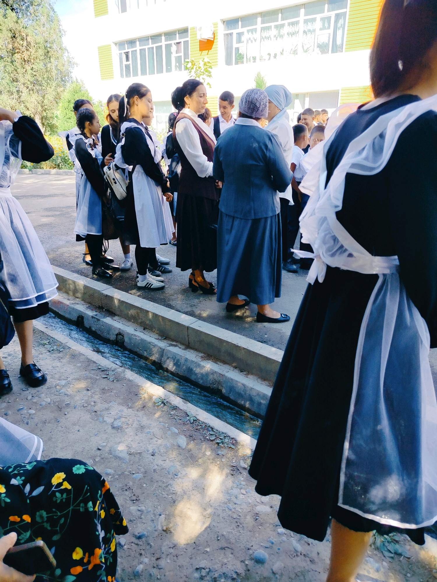 The image depicts a group of individuals, likely students, standing outdoors in front of a building. Many are wearing school uniforms consisting of black dresses with white aprons or collars. Some students are facing the camera, while others are looking away or interacting with each other. In the foreground, a person's hand is visible holding a smartphone. The ground is paved with gravel and there is a concrete drainage ditch running alongside the students. The background shows a building with yellow and green accents and windows. The lighting suggests it is daytime. The context provided indicates the location is the city of Kölkent, Kazakhstan.