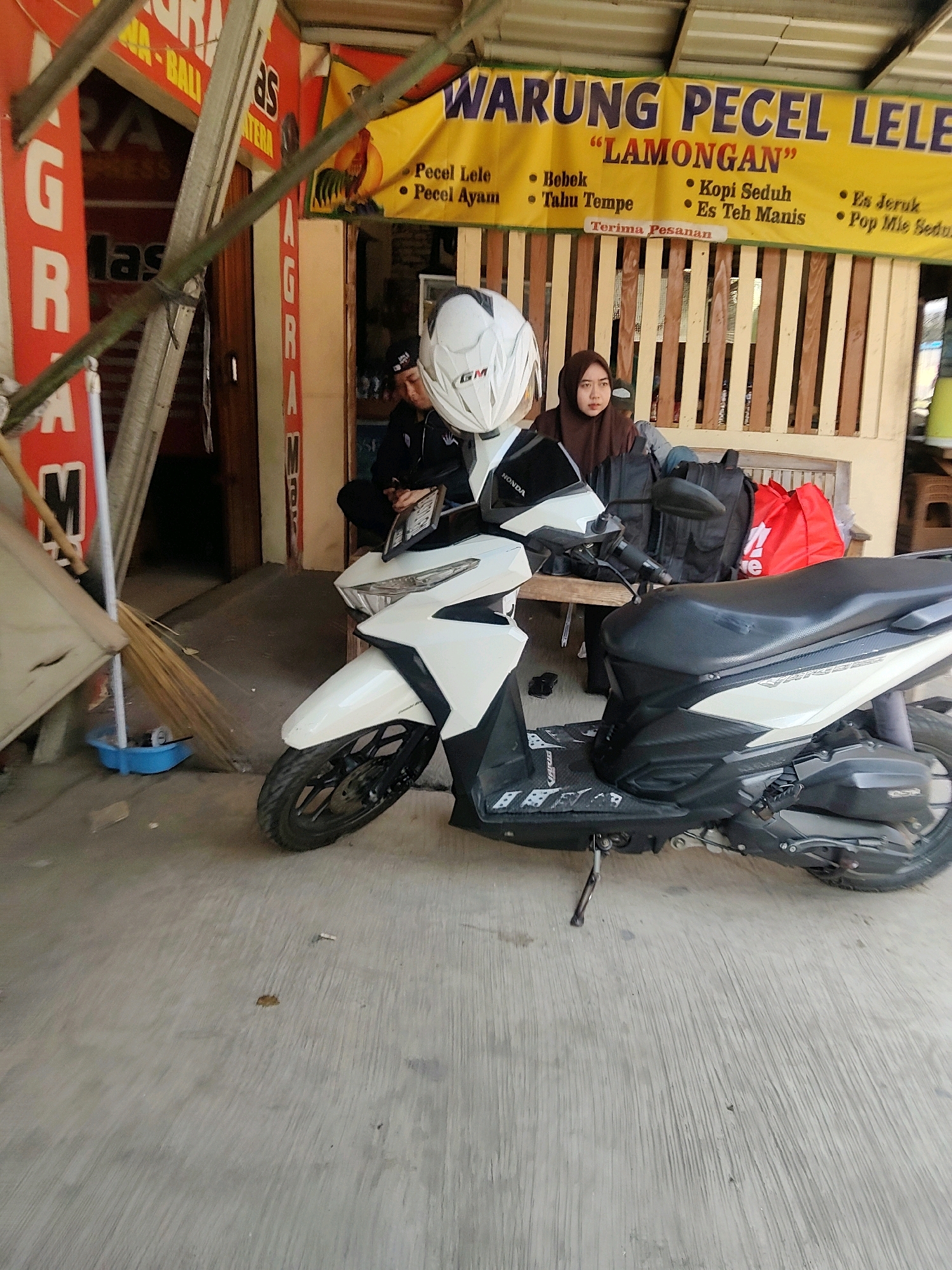 The image depicts a scene outside a roadside food stall in Sukaresmi, Indonesia. In the foreground, a white and black scooter is parked. Behind the scooter, two individuals are seated on a bench. One is a person wearing a black jacket and a dark beanie, looking down at their hands, holding a mobile phone. Next to them, a woman wearing a brown hijab and a dark jacket is looking towards the right of the frame. Behind them, a yellow banner with blue text advertises "WARUNG PECEL LELE LAMONGAN" and lists menu items such as "Pecel Lele", "Bebek", "Tahu Tempe", "Kopi Seduh", "Es Teh Manis", "Es Jeruk", and "Pop Mie Seduh". Red bags and black backpacks are also visible on the bench. To the left of the scooter, a broom is leaning against a blue bucket, with a broom of straw partially visible. Red signage with white text reading "GRAM" is also visible on the left side of the image, partially obscured by bamboo structures. The ground is paved with concrete.