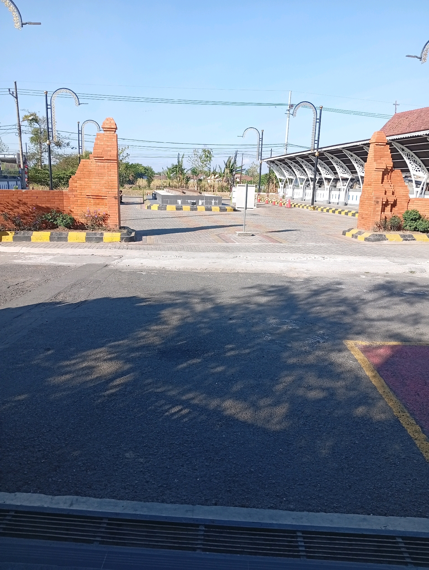 A clear sky with a few wispy clouds is visible above a rural landscape in Tebon, Indonesia. In the foreground, a paved road with a shadow of tree branches casts across its surface leads to a brick archway. Beyond the archway, a covered seating area with a red tiled roof and ornate white pillars extends into the distance. The area is marked with yellow and black striped bollards and a few traffic cones. A person can be seen standing near the archway, and a small bush with pink flowers grows at its base. The overall scene suggests a public space, possibly a rest area or a waiting area, given the covered seating and road infrastructure.