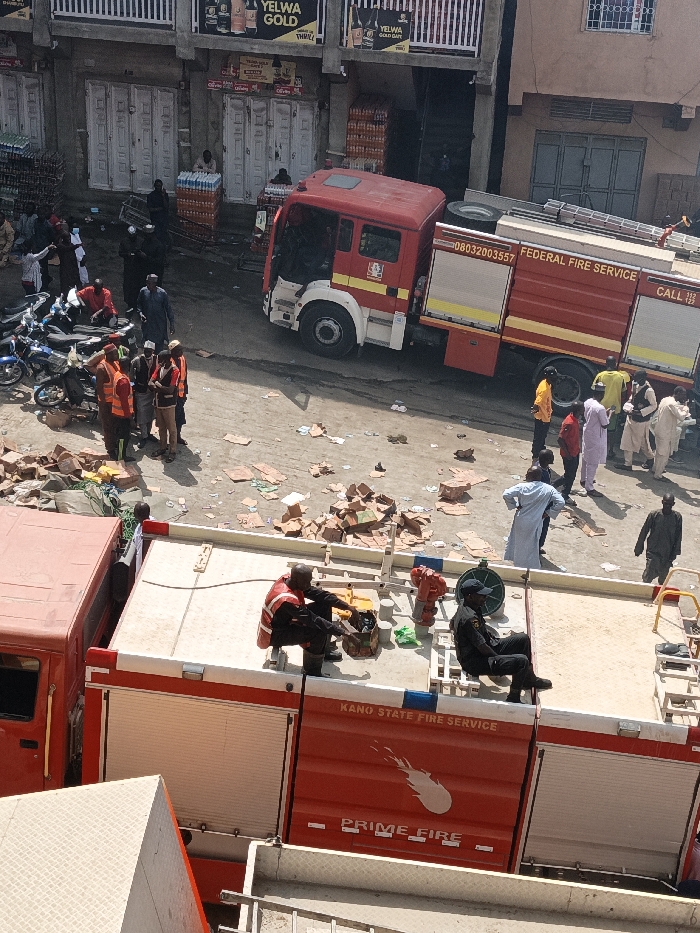 An overhead view in Fagge C, Nigeria, depicts an outdoor scene dominated by emergency vehicles and personnel. Two red fire trucks are present: a large Federal Fire Service truck (bearing number 08032003357 and "CALL 112") is parked diagonally on a dirt surface, and a Kano State Fire Service "PRIME FIRE" truck is partially visible in the foreground. On the roof of the foreground truck, one individual in a red vest kneels, seemingly attending to a red hydrant connection and associated equipment, while another person in dark attire sits nearby.
The ground is uneven and extensively littered with scattered cardboard boxes, papers, and other refuse. Approximately 20-30 individuals are observed throughout the scene. Several men wearing orange or red high-visibility vests, identifiable as emergency responders, are grouped near the Federal Fire Service truck and a cluster of parked motorcycles on the left. Other individuals, dressed in various civilian clothes, including traditional long robes, stand in small groups or walk across the area, observing the scene. In the background, two multi-story buildings feature ground-floor shops, one displaying "YELWA GOLD" signage, with stacks of bottled beverages visible outside. The lighting indicates bright daylight. No active fire is apparent; the scene suggests a post-incident or standby situation.