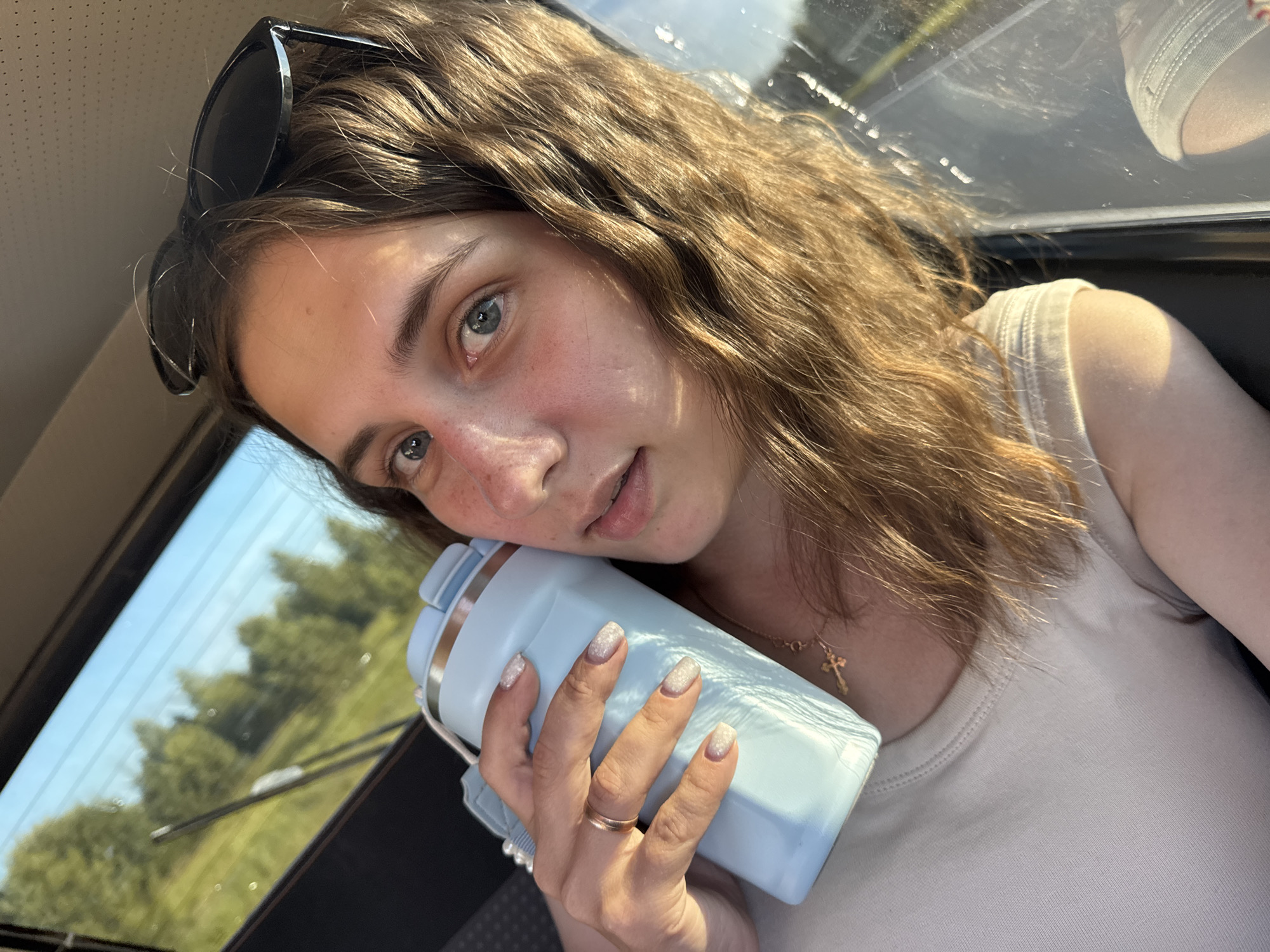 A young woman with light brown hair and blue eyes is holding a light blue tumbler in her right hand. She is wearing a beige t-shirt and a gold cross necklace. Sunglasses are perched on top of her head. The background shows trees and a bright sky, suggesting an outdoor or travel setting. The image appears to be a selfie taken inside a vehicle, possibly a car, as evidenced by the reflection in the window and the interior details. The location is identified as Vakhrino, Russia.