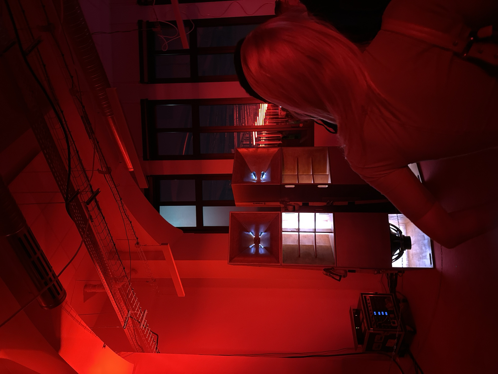 This image depicts an indoor scene, likely a performance venue, club, or art installation in Paris, France, dominated by an intense red light. The perspective is tilted, looking upwards from below, giving a dynamic and immersive feel.
In the foreground, the back of a person's head with bright blonde or light-colored hair is visible, along with part of their shoulder and arm. They appear to be looking down or away from the camera, observing the space.
The environment is characterized by industrial elements and audio equipment. Several large speakers are prominent. One horn-shaped speaker emits a distinct blue, cross-like glow from within, while an adjacent speaker unit features a light-colored grille with vertical dividers. Further to the right and lower, a large speaker cone is partially visible. Below this, a rugged electronic equipment rack case with multiple blue indicator lights hangs suspended by chains, connected by various cables.
The ceiling and walls feature exposed infrastructure, including metal grates or cable trays housing numerous wires and pipes, suggesting a utilitarian or industrial design. Windows with dark panes are visible in the background, some reflecting ambient light. The pervasive red lighting creates a dramatic and energetic atmosphere, hinting at an evening or night event. No discernible text is visible in the image.