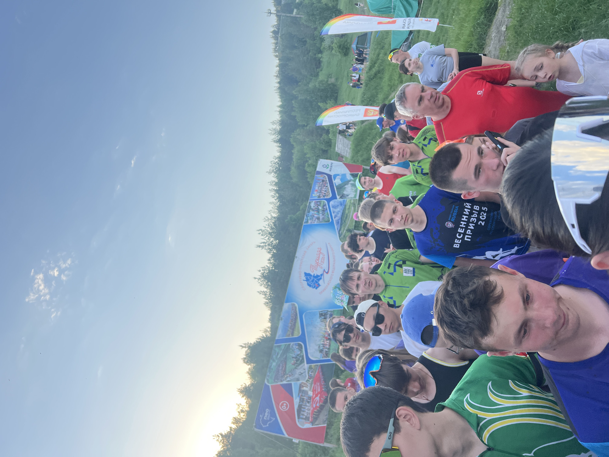 A crowd of people, primarily young men, are gathered in an outdoor location, presumably in Khvalynsk, Russia. Many are wearing athletic clothing, suggesting a sporting event or competition. A large banner with the words "Весенний Призыв 2025" (Spring Conscription 2025) and "Russia" is visible. Other signage and banners with logos are also present. The environment includes trees and grass. The sky is visible. Some people are looking towards the camera, while others are engaged in conversation or looking in different directions. The photo seems to capture a moment of anticipation or waiting.