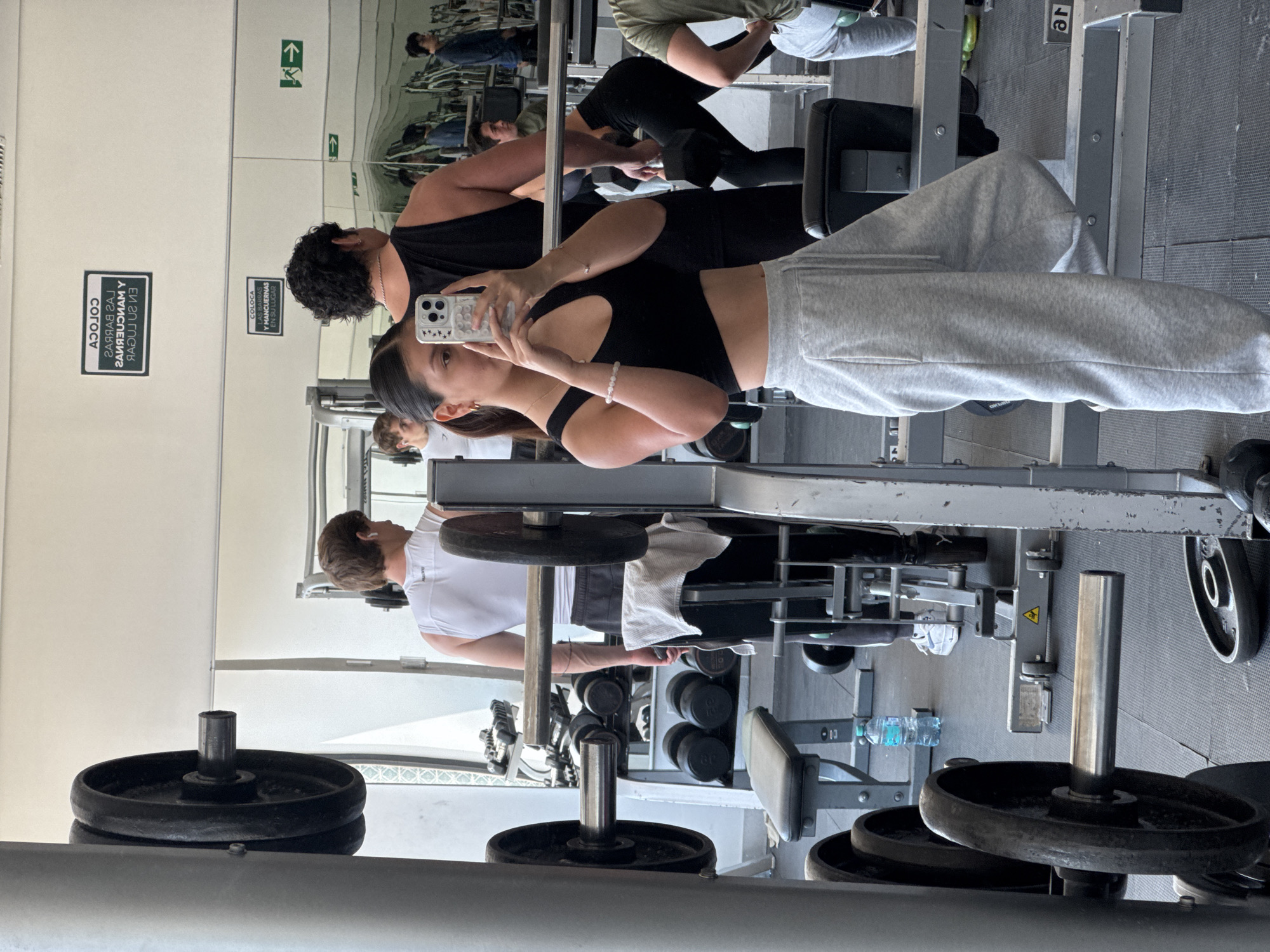 The image shows a gym, presumably in San Andrés Cholula, Mexico. A woman in a black top and grey sweatpants is taking a picture of herself in a mirror. Behind her, a man with dark curly hair is also exercising. Other gym-goers and weight equipment are visible in the background. The environment appears to be a well-lit indoor space with typical gym equipment such as weight machines, dumbbells, and weight plates. There are informational signs on the wall. The gym seems relatively busy. A bottle of water is visible next to some gym equipment.