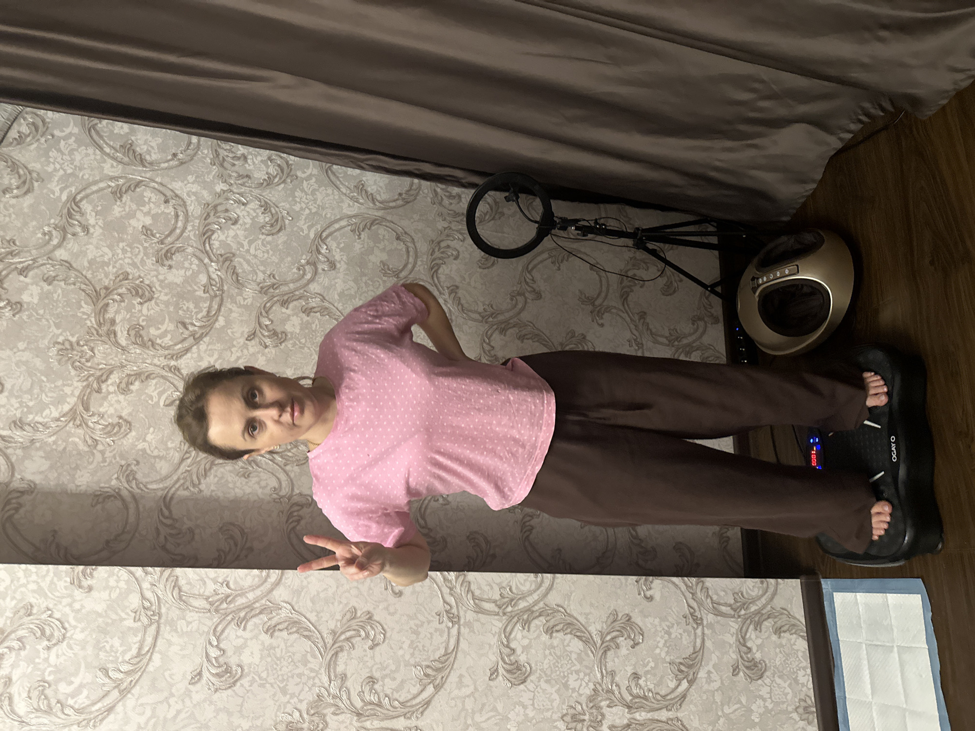 A woman stands on a black vibration plate, a common piece of fitness equipment, in a room with ornate wallpaper. She is wearing a pink t-shirt with white polka dots and brown sweatpants. Her left hand is on her hip, and her right hand is raised in a peace sign. To her right, a ring light on a tripod is visible, and further to the right is a large brown curtain. In front of the vibration plate, to the left, a white and blue padded mat is on the floor. A golden foot massager is positioned on the floor to the right of the vibration plate. The scene appears to be indoors, likely in a residential setting. The presence of the vibration plate and foot massager suggests a focus on personal health and wellness. The wallpaper and curtains indicate a decorative interior design. The lighting is dim, creating shadows that add to the atmosphere of the room. The overall impression is of a personal space dedicated to self-care routines. The location is identified as Verkhniy Yurt, Russia.