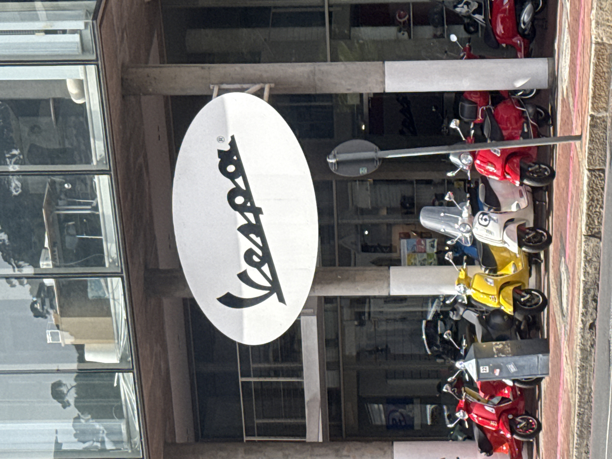 An exterior, eye-level photograph captures the front facade of the Vespa dealership located at 145 Buitengracht Street, Cape Town, South Africa, under bright daylight conditions. The building features a multi-story structure with extensive glass windows on both the ground and upper floors. A large, oval-shaped white sign with the black "Vespa" logo is prominently mounted above the ground floor entrance, extending between two concrete support pillars.
On the ground level, several Vespa scooters are parked in a line on the paved sidewalk in front of the glass-fronted showroom. These scooters are predominantly red, yellow, and white, with at least one dark grey/black model also visible. Specific scooters include a white model with the number "6" on its front shield and multiple red scooters.
On the sidewalk, a grey street utility box with the number "22" is positioned to the left of a dark grey, circular signpost. Reflections in the upper-story windows reveal an interior office setting with a desk and a faint silhouette of a person seated on the left side. The foreground shows a paved sidewalk transitioning to a street curb and asphalt road.