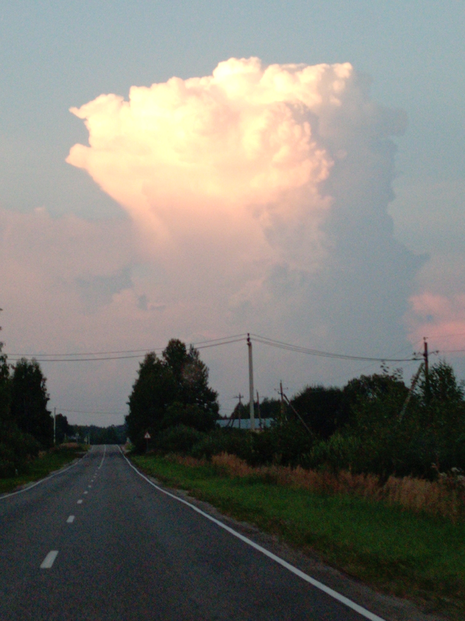 The image depicts a rural road leading towards a horizon under a dramatic sky. A large, fluffy cloud formation, tinged with pink and yellow from the setting sun, dominates the upper portion of the frame. The road is paved and marked with white lines, suggesting it is a secondary route. On either side of the road, there is green vegetation and scattered trees. In the distance, power lines are visible, supported by poles, indicating some level of infrastructure in the area. The overall atmosphere is serene and evokes a sense of rural tranquility at dusk. The image does not contain any people or specific identifiable buildings that would pinpoint a location within Leskovo, Russia, beyond the general assumption of a rural setting consistent with the provided metadata.
