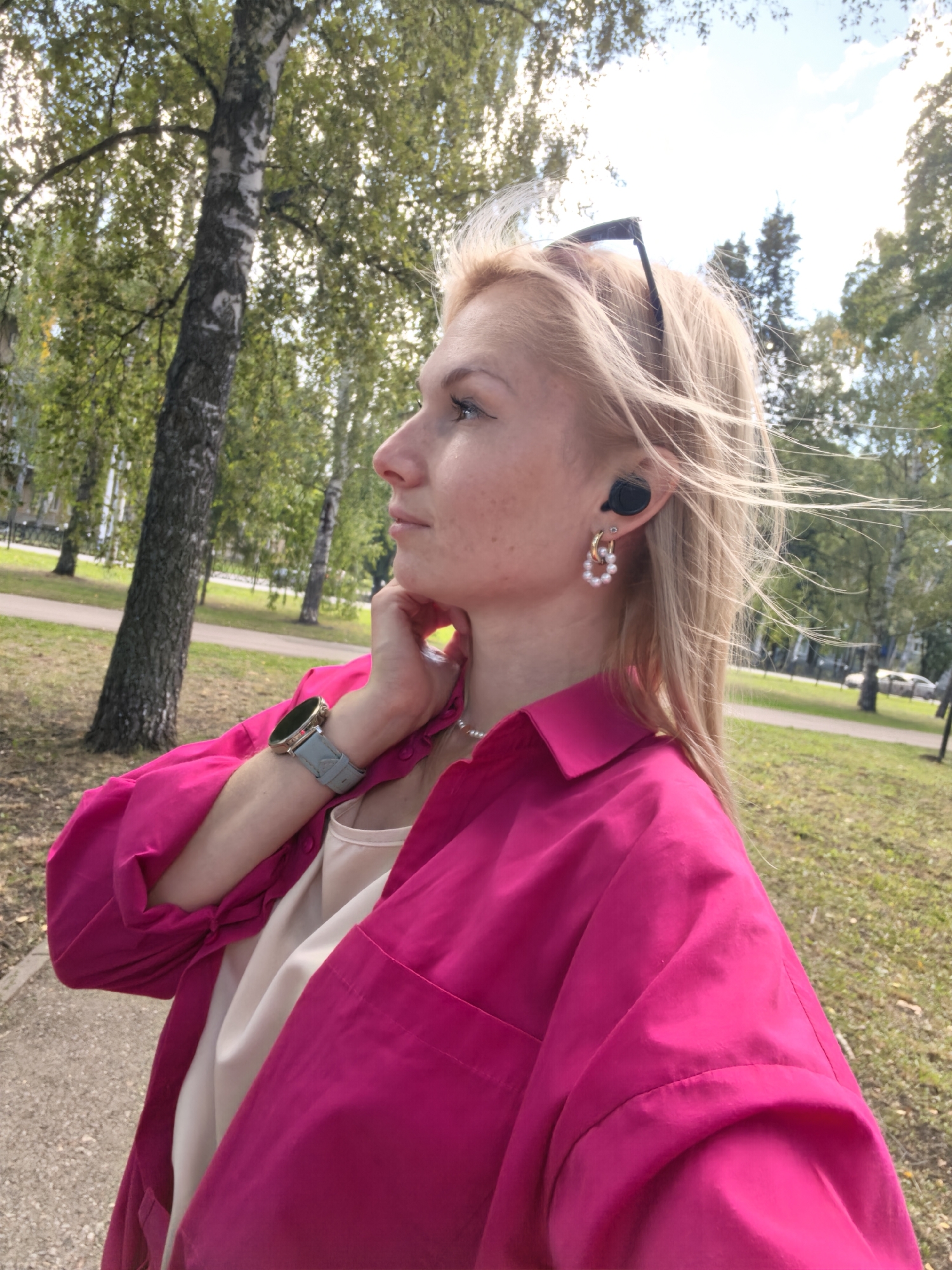 The image shows a blonde woman from the chest up, looking to her left. She is wearing a bright pink collared shirt over a white top. She has earrings with pearls and a watch on her left wrist. Black earbuds are in her ears. She is outdoors in a park-like setting with trees and grass. The lighting suggests it is daytime, with sunlight filtering through the trees. The presence of birch trees and the general landscape are consistent with areas found in Sterlitamak, Russia.