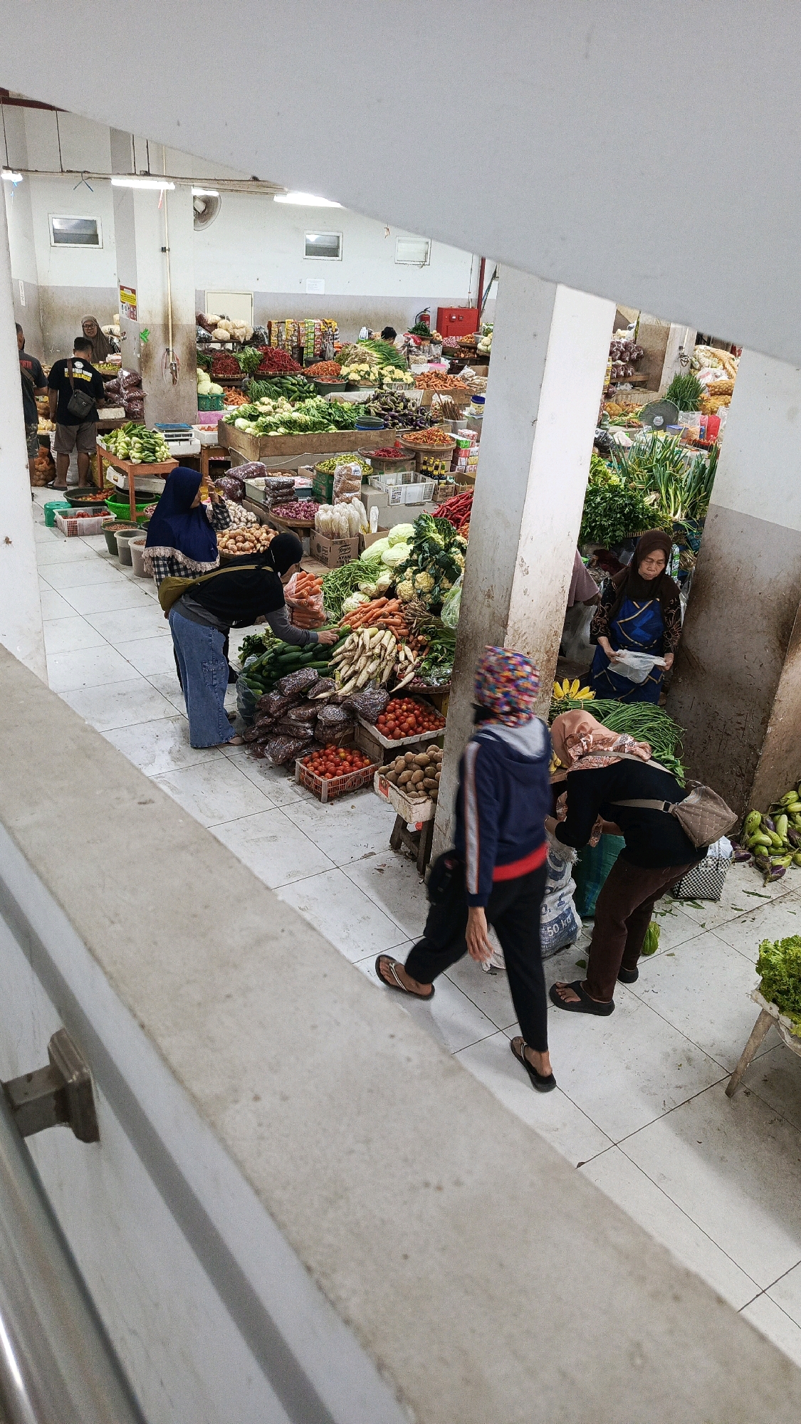 The image depicts a bustling indoor market in Nologaten, Indonesia, likely a vegetable market given the abundance of produce. The scene is viewed from an elevated perspective, possibly a balcony or upper level, looking down onto stalls laden with fresh vegetables. Several individuals are present, engaged in shopping and selling activities.
In the foreground, a person wearing a blue jacket and a patterned headscarf is walking away from the camera, their legs and feet clearly visible. To their right, another person in a dark top and brown pants is bending down, possibly examining or purchasing items. Further to the left, two women are interacting at a stall; one is wearing a blue hijab and appears to be holding produce, while the other, in a black top and blue pants, is reaching towards the vegetables.
The stalls are filled with a variety of colorful vegetables, including leafy greens, tomatoes, cucumbers, carrots, onions, and potatoes, among others. The market floor is tiled, and the stalls are constructed from wood and metal. Lighting appears to be from overhead fixtures, illuminating the scene. The overall impression is one of a busy, active marketplace where commerce and daily life converge.