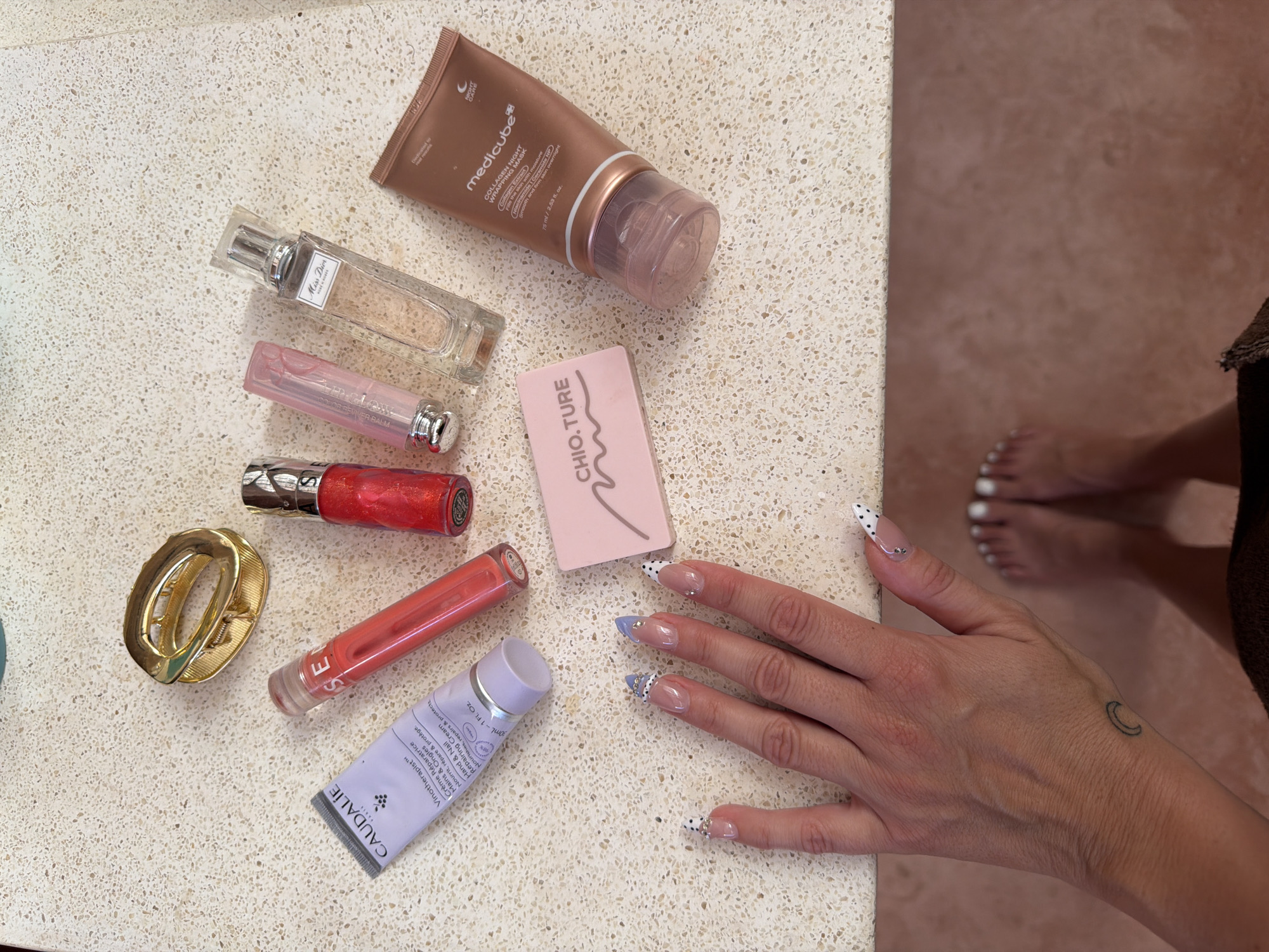 The image shows a flat lay of various beauty products on a speckled countertop, next to a person's hand and feet. The beauty products include a brown tube labeled "medicube Collagen Night Wrapping Mask", a clear perfume bottle, a pink Dior lip balm, a red lip gloss, a gold hair clip, a coral lip gloss with "SE" visible on the tube, a lavender tube labeled "CAUDALIE Vinotherapist Hand & Nail Repairing Cream", and a pink Chioture product compact. The person's hand is resting on the countertop, with a small crescent moon tattoo visible on the wrist and decorative nail art. The person's bare feet are partially visible on the right edge of the image. The location is indeterminate, with no clues as to it being North Kuta, Indonesia.