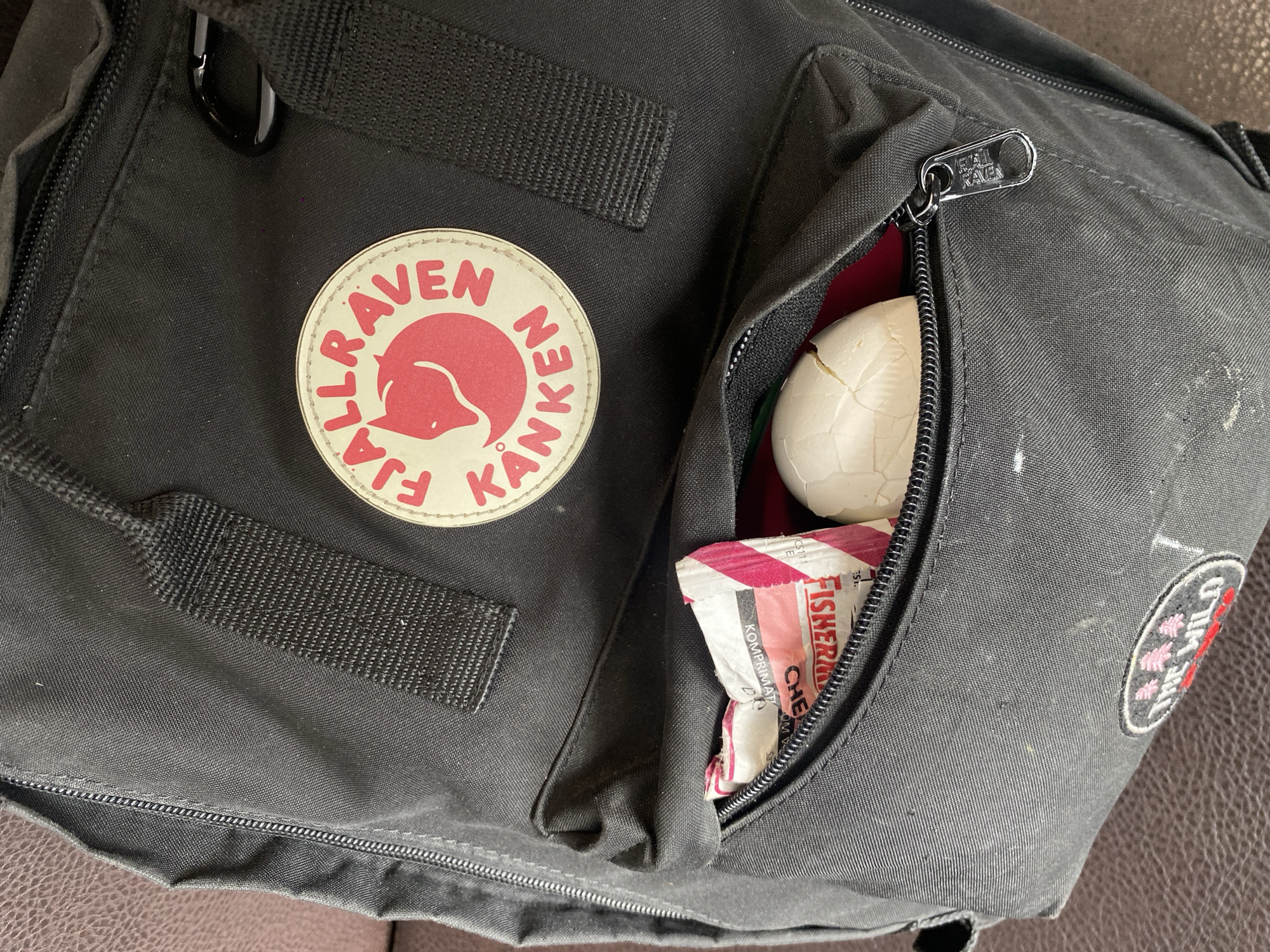 The image depicts a black Fjallraven Kanken backpack. The backpack features the brand's circular logo with a red fox design on one of the front pockets, which also has several straps. The zipper of another pocket is partially open, revealing the edge of a red interior, a cracked white eggshell, and a partially visible package of "Fisherman's Friend Cherry Komprimaten" lozenges. There is an embroidered patch on the side of the bag that reads "THE WILD". The bag rests on a brown surface.