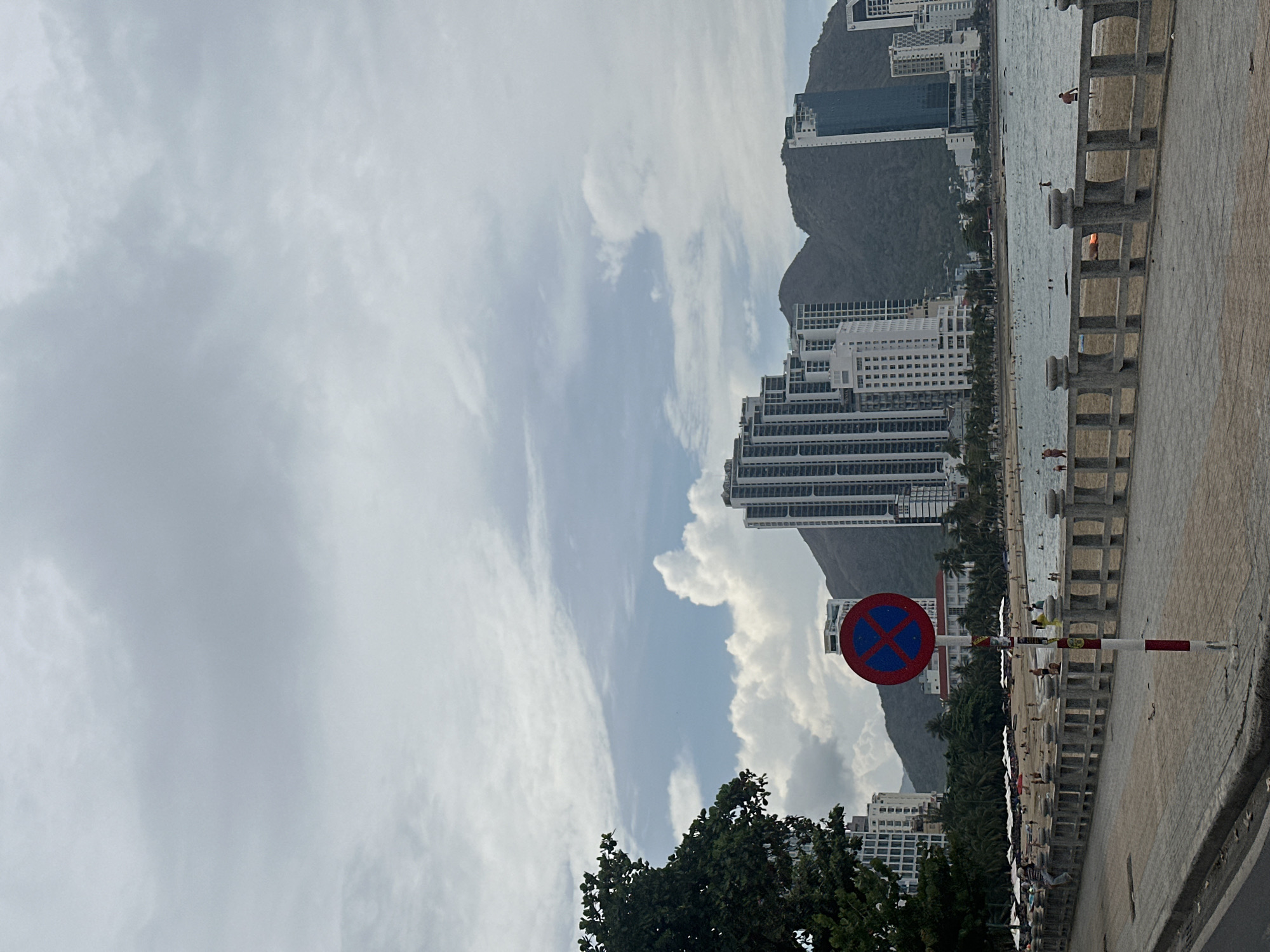 The image depicts a view of Nha Trang, Vietnam, specifically within the Bac Nha Trang Ward. The scene includes a view from a street or sidewalk along the coast, evidenced by a concrete barrier along the right edge of the image. In the background, there is a beach area with people, the ocean, and a row of high-rise buildings against the backdrop of mountains. A no parking road sign is in the foreground. The sky is overcast with visible cloud formations.