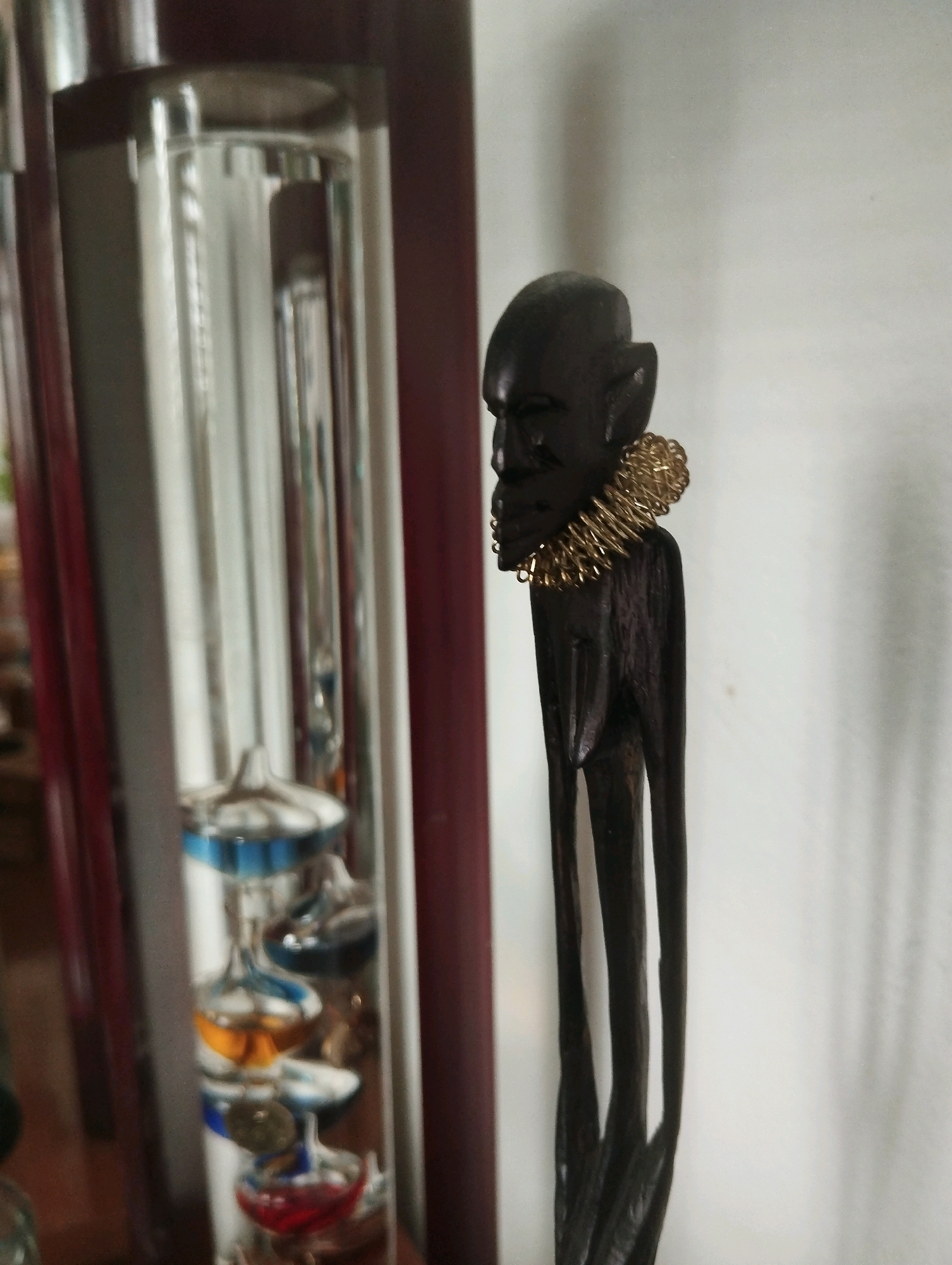 The image shows a close-up of two objects against a white wall. On the left is a Galileo thermometer, a glass tube filled with colored liquid and small glass spheres, each marked with a different temperature. The thermometer is housed in a clear glass cylinder, which is itself enclosed in a dark red frame. To the right of the thermometer is a dark, carved wooden figurine of a person with long, thin legs and a detailed face. The figurine is wearing a coiled, gold-colored necklace around its neck. The scene is indoors, likely in a residential setting. The lighting is soft, casting a shadow of the figurine on the wall to its left. There is no indication of the specific location within Ashkelon, Israel, beyond it being an indoor environment.
