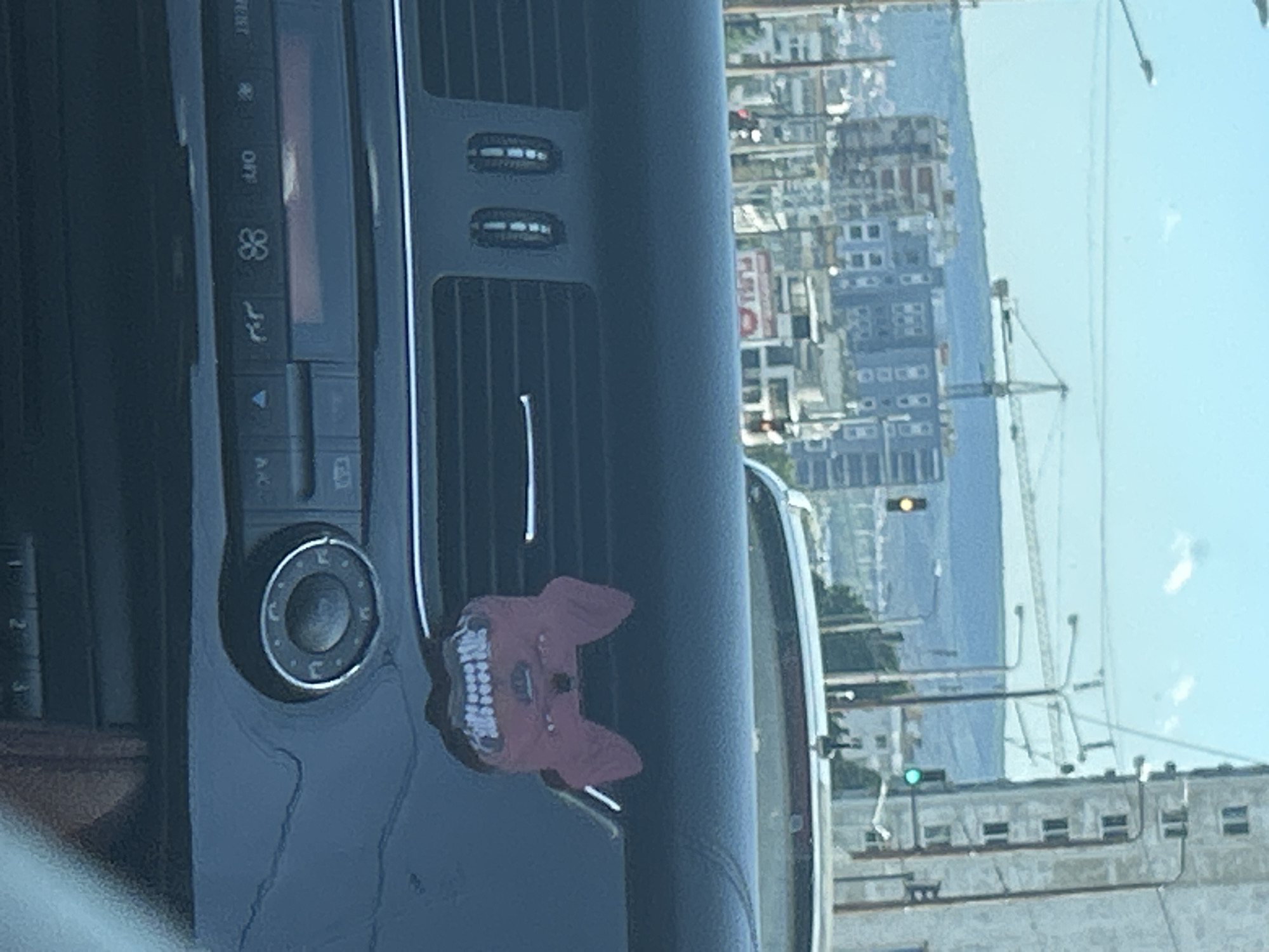 The image shows a close-up view of a car's interior dashboard with a vntilation grille and various controls. Attached to the grille is a red air freshener shaped like a stylized, monstrous dog or wolf head with large teeth and wide eyes. In the background, through the car's windshield, a cityscape is visible. This includes several multi-story buildings, some under construction with cranes, traffic lights, and a hill in the distance. The sky is bright blue with a few clouds. The scene appears to be an urban environment, likely Varna, Bulgaria, given the context provided.