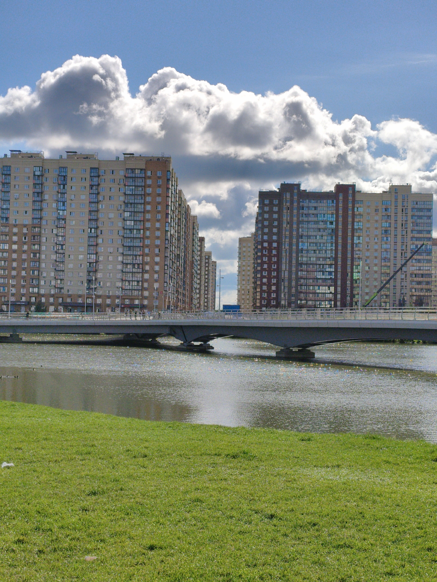 An eye-level, vertical, medium shot captures a waterside view of a modern city, with a focus on apartment buildings and a bridge. In the foreground, a lush green lawn slopes gently towards the water. The water itself is calm with a shimmering surface, reflecting the bright daylight. Two dark shapes, possibly ducks, are visible on the water. Spanning the scene is a modern bridge with a gray concrete deck and white metal railings. The bridge supports are dark gray. Behind the bridge, a series of tall, multi-story apartment buildings dominate the background. These buildings vary in color, with some appearing beige and others a reddish-brown or gray. Many have numerous windows and balconies. Above the buildings, a vast expanse of blue sky is filled with dramatic, puffy white clouds. The lighting suggests it is daytime, with the sun casting bright light and creating reflections on the water. The overall impression is of a contemporary urban landscape by the water on a clear day with dynamic cloud formations.