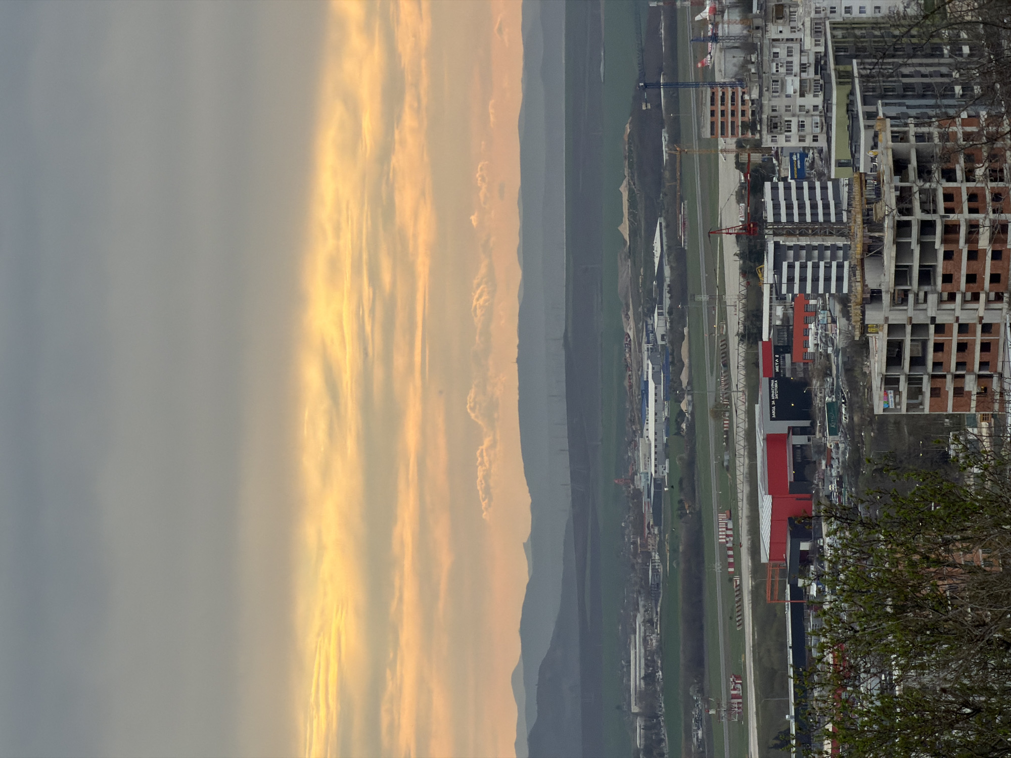 The image depicts an aerial view of a cityscape, likely Varna, Bulgaria, given the architecture and landscape. In the background, a mountain range is visible, transitioning into a valley area. A runway stretches from the background into the mid-ground of the image, suggesting the presence of an airport or airbase. Nearer to the foreground, buildings and infrastructure projects are under construction with numerous cranes visible. The construction area is densely populated with high-rise buildings and residential complexes of varying architectural styles. The sky is overcast with a mix of light and dark clouds, suggesting either sunrise or sunset. The foreground contains trees.