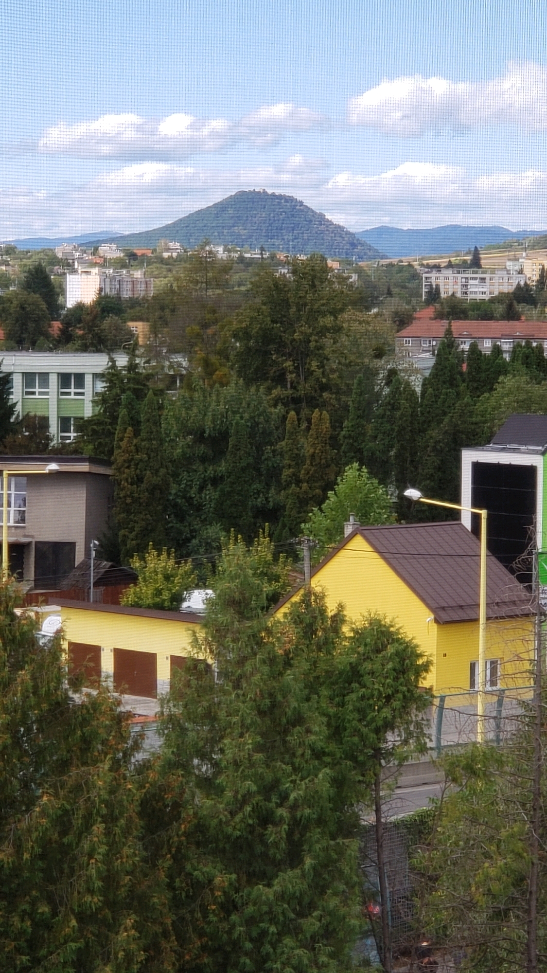 The image depicts a view of the city of Prešov, Slovakia, with a prominent conical mountain in the background. The foreground features residential buildings, including a yellow house with a brown roof and a building with garage doors. Several trees, some appearing to be cypress or similar evergreens, are scattered throughout the scene. A street light with a yellow pole stands near the yellow house. In the mid-ground, there are other buildings, including a multi-story white building with green trim, and a smaller grey building. The background is dominated by the conical mountain, partially covered in trees, and a hazy sky with scattered clouds. The overall impression is of a residential area with a natural landscape feature.