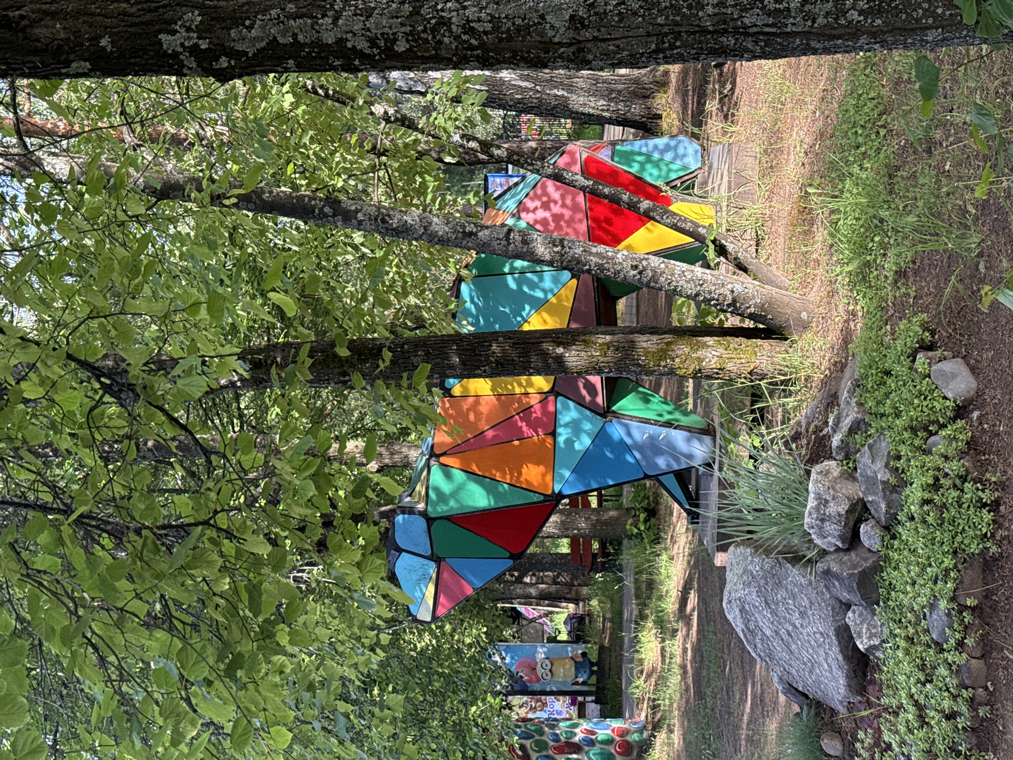 The image presents a scene at the Lukomorye recreation park in the city of Chaikovsky, Perm Krai, Russia. A colorful, geometric sculpture of what appears to be a bear or similar animal is prominently displayed in a wooded area. The sculpture is composed of numerous triangular and quadrilateral pieces of metal, painted in bright colors such as red, blue, yellow, green, and orange. The sculpture is positioned amidst trees with green foliage and dappled sunlight. The trunks of the trees have gray bark with some lichen growth. The ground cover includes grass, small plants, and a rocky area on the right side of the frame. In the background, a pathway or clearing can be seen with more colorful structures and signs, indicating a recreational or playground area. The image is taken from a low angle, with trees framing the top and sides of the view.