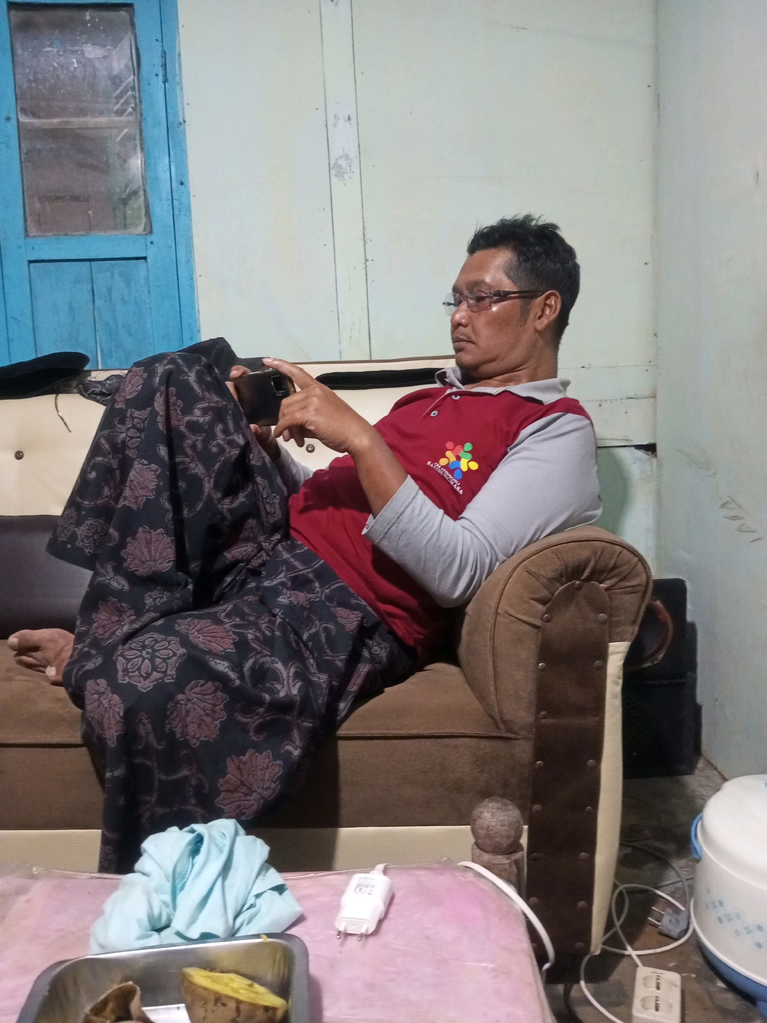 A man with short dark hair and glasses is sitting on a brown sofa and looking at his phone. He is wearing a red polo shirt with grey sleeves and a patterned sarong. The room appears to be a home in Andongsili, Indonesia, with a blue wooden door and window on the left and a pale green wall on the right. In the foreground, a tray with cooked sweet potatoes and a folded light blue cloth are visible, along with electrical cords and outlets.