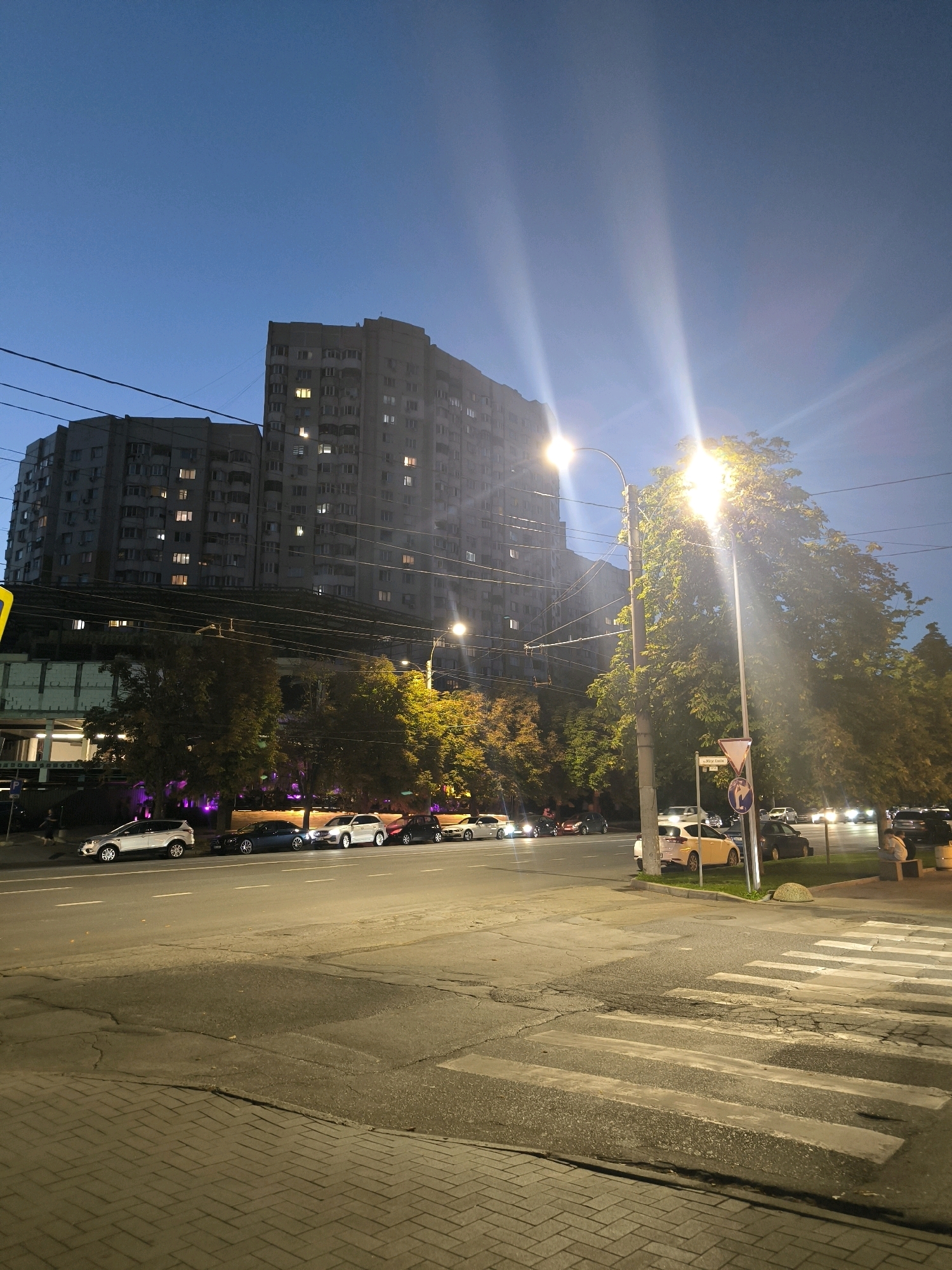 The image depicts an urban street scene in Chișinău, Moldova, during twilight. Two large, multi-story apartment buildings dominate the background, their windows illuminated. A streetlamp with a bright light source illuminates the foreground and a mature tree to its right. The street itself is paved, with a crosswalk visible. Several cars are parked along the side of the road and in motion further down. A sidewalk made of brick pavers runs along the left side of the image. The sky is a deep blue, indicating the transition from day to night.
