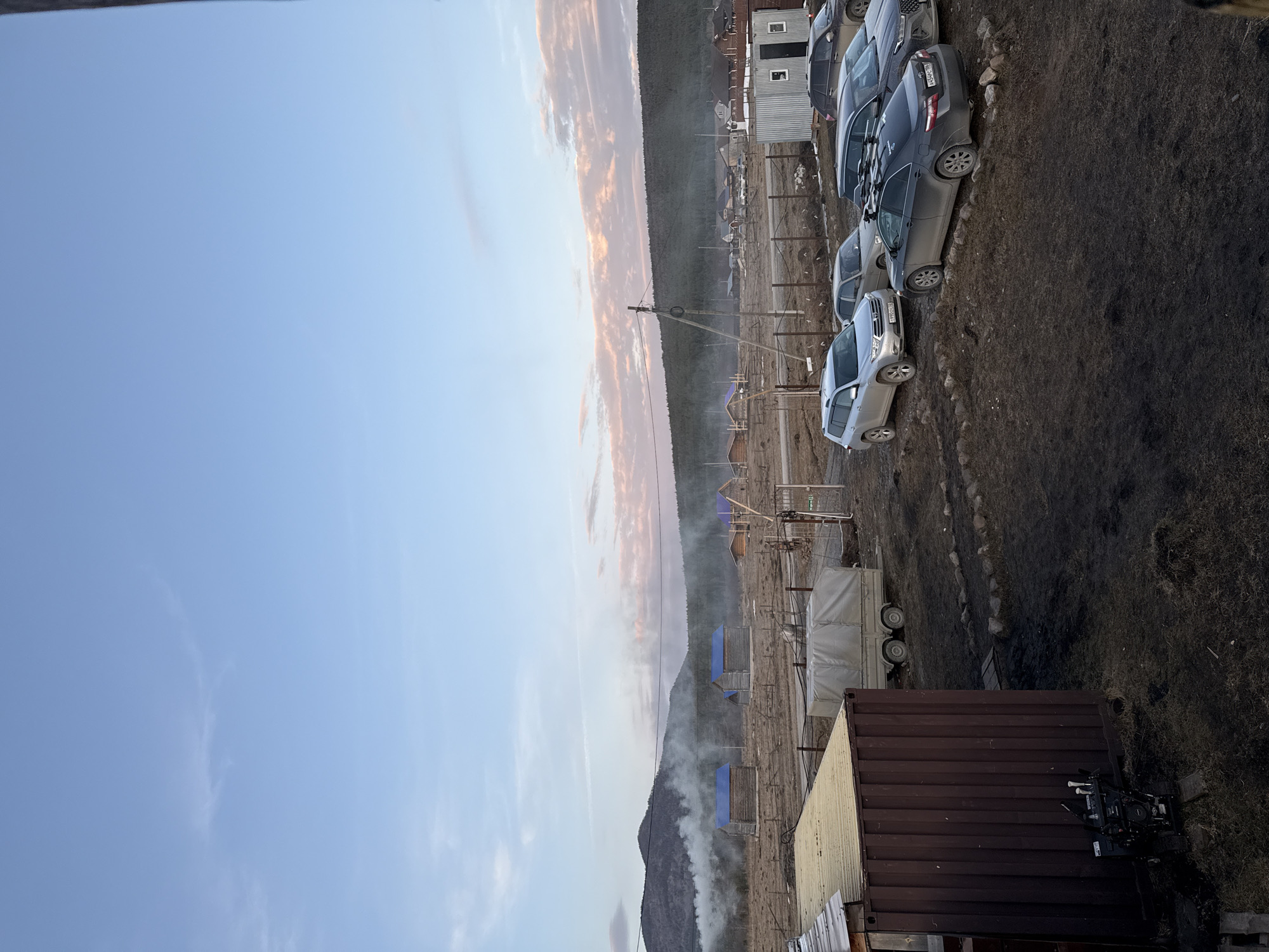 This image depicts a rural or semi-urban landscape in the city of Tyulyuk, Russia, under what appears to be late afternoon or early evening lighting conditions. The sky is predominantly light blue with scattered wispy clouds, some displaying pinkish hues towards the horizon on the right.
In the immediate foreground on the left, a portion of a dark brown corrugated shipping container or similar structure with a flat, light tan roof is visible. Parked adjacent to this structure, resting on a wooden platform or pallet, is a small, black tracked utility machine featuring white components on its upper section. The ground in the foreground is uneven, consisting of dark, disturbed earth with sparse, dry vegetation.
The midground features an unpaved parking area on similarly uneven, dark ground. Several vehicles are parked: a silver Toyota Highlander SUV (license plate T900TO 102), a dark gray sedan (partially obscured), and a dark gray Toyota Camry sedan equipped with roof racks (license plate X454CC 196). A dark SUV is partially visible on the far right. Further back in the midground, a light-colored, two-axle utility trailer with a white tarp cover is parked. A simple metal fence line with a metal gate on the right traverses the midground. A wooden utility pole with overhead wires is centrally located on the right side of the frame. To the right of the parked vehicles, a small, rectangular metallic shed with two dark doors and two small windows is present.
The background to the left and center reveals an open, barren field dotted with multiple small, rustic log cabins or wooden structures, each distinguished by a bright blue corrugated roof. White plumes of smoke are actively rising from at least two locations within this field, suggesting combustion. Behind the field, a dark, tree-covered mountain range extends, partially veiled by the rising smoke. Towards the background on the right, a dense forest line on a sloping hill is visible, with additional structures indicative of a village appearing further in the distance. No individuals are visible in the scene.