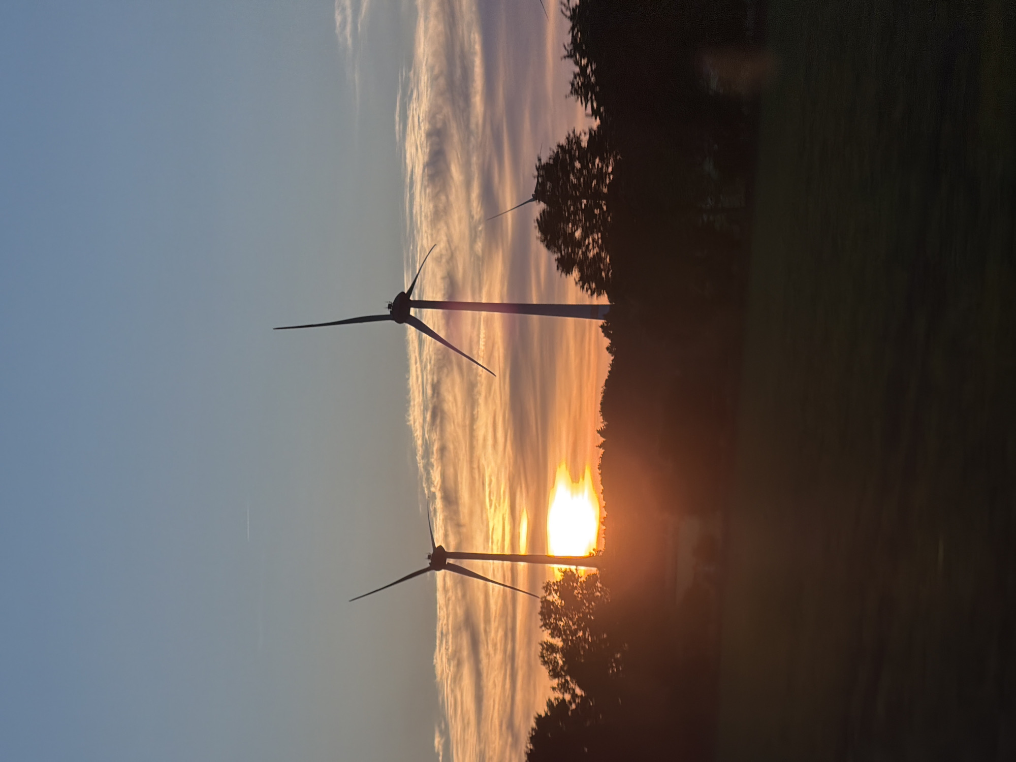 The image depicts a sunset with two wind turbines in the foreground. The turbines are silhouetted against the bright setting sun, which is partially obscured by trees and clouds. The sky is a mix of blue and orange hues, and there are cirrus clouds above. The photo was likely taken near Harsefeld, Germany, because that is where the wind turbines are located.