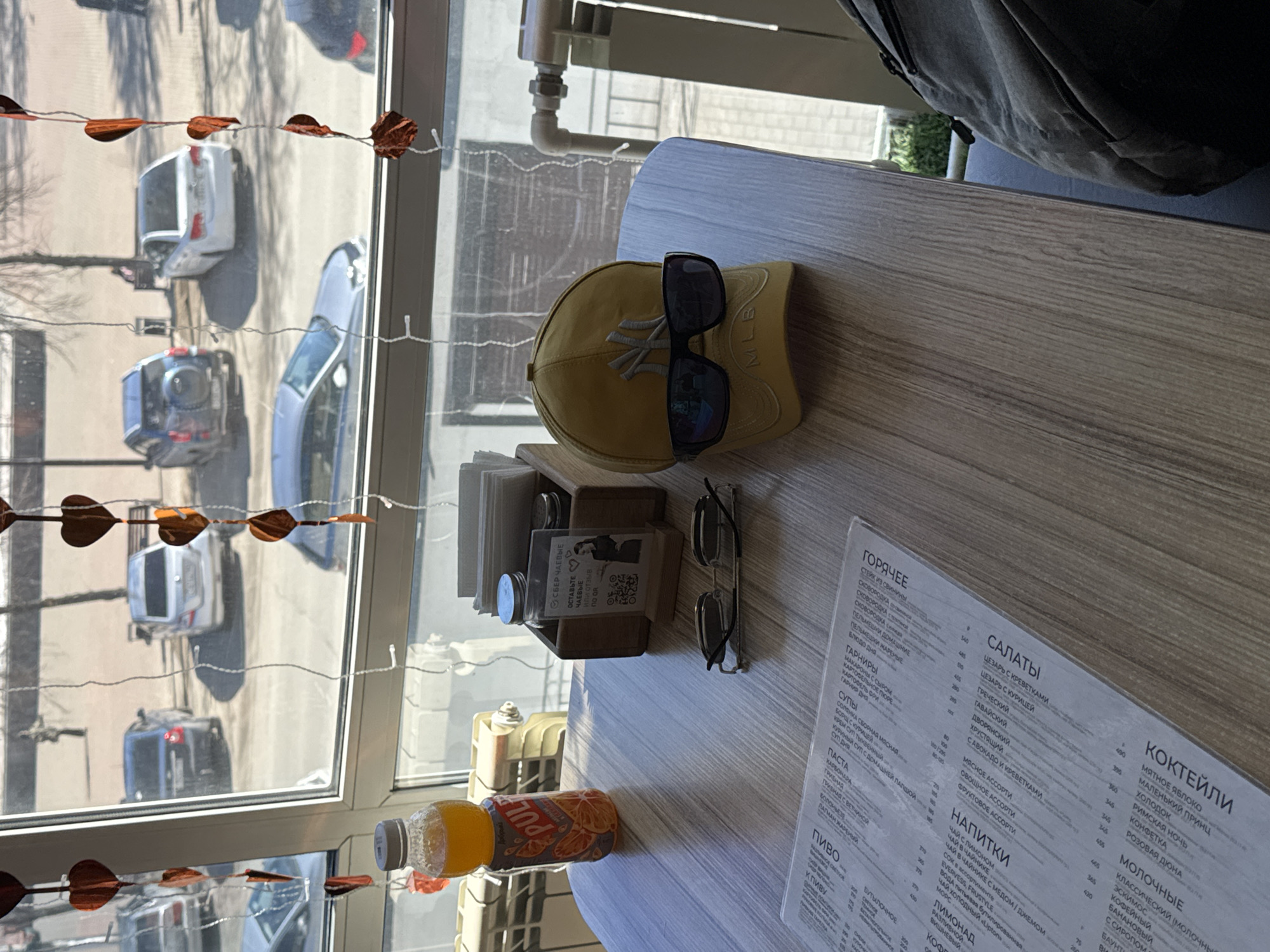 The image depicts a table in a cafe or restaurant. A menu, a yellow New York Yankees baseball cap with sunglasses resting on it, a juice bottle, napkins, and a pair of eyeglasses are on the table. A bag hangs on the chair behind the table. A window reveals a street scene in Chaikovsky, Russia. Several cars are visible on the street. The location is likely a business that serves food and drinks in Chaikovsky, Russia.
