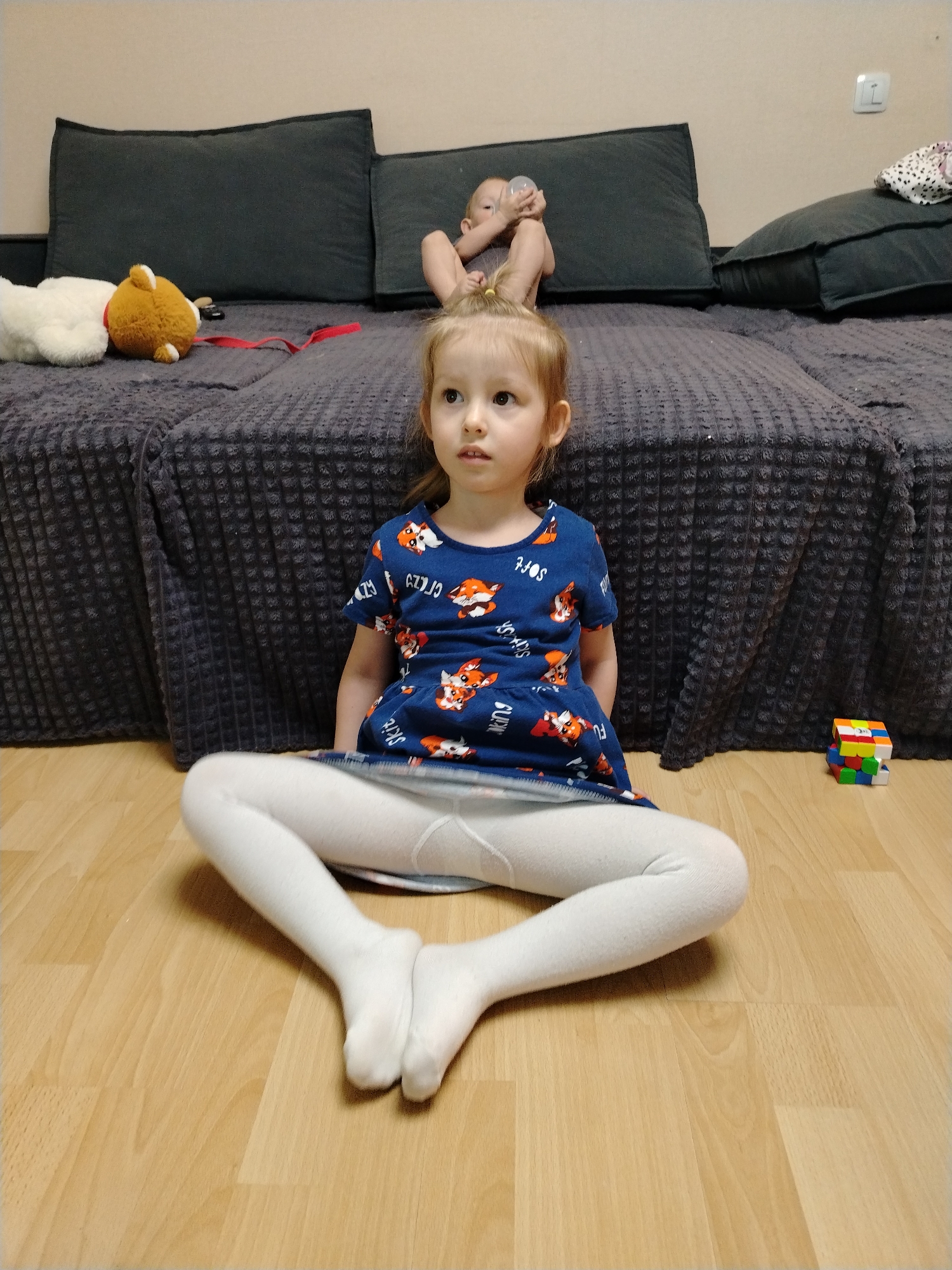 A full, eye-level shot displays a young girl seated cross-legged on a light wood floor. She wears a blue dress with a fox pattern and white tights. Behind her, a large, grey textured couch extends across the frame, upon which a baby lies on their back, holding a bottle. To the girl's right, a Rubik's cube sits on the floor. A stuffed animal and other toys are visible on the couch. The photo was taken indoors in a room located in Chaikovsky, Russia.
