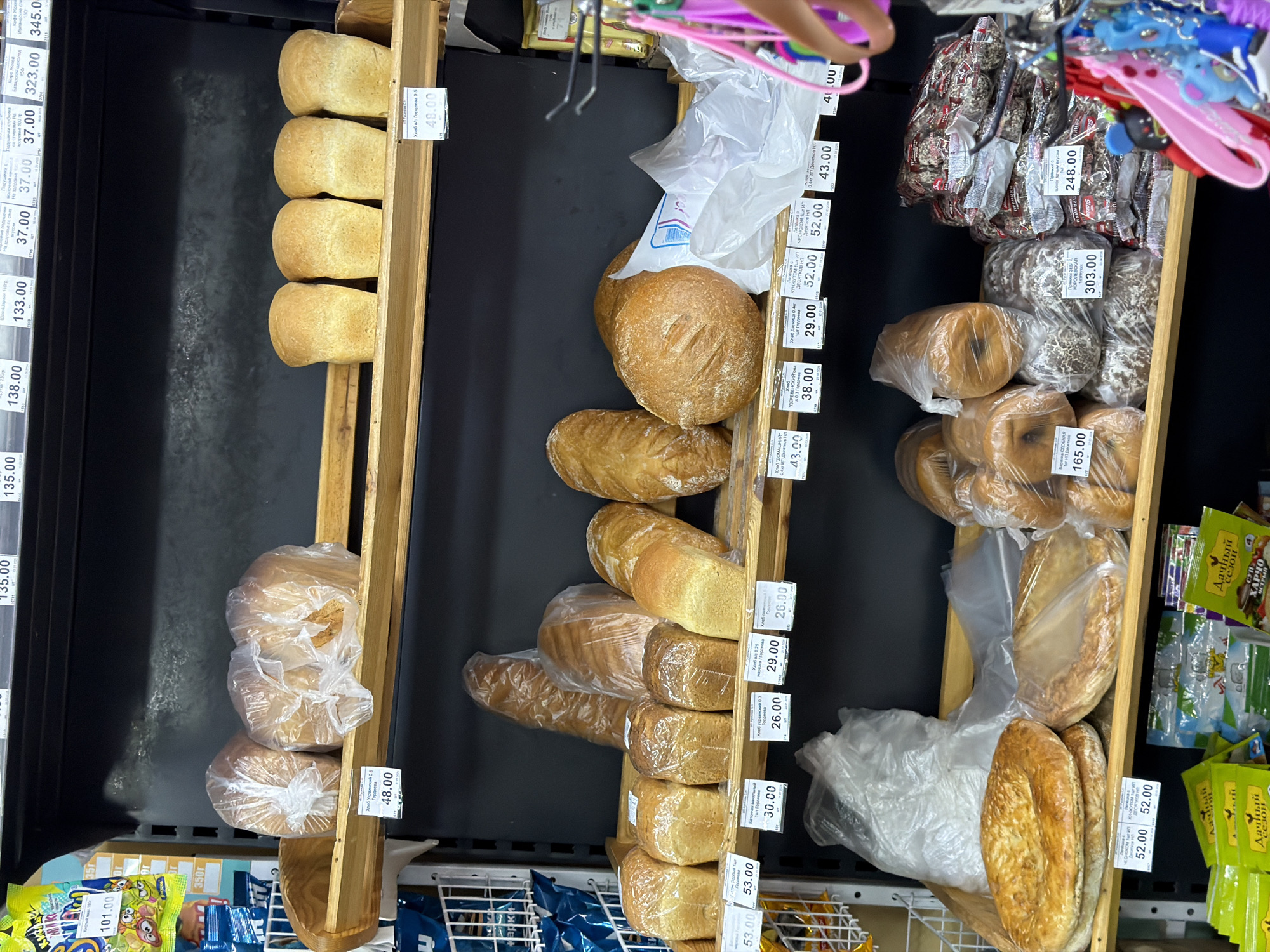 The image shows a shelf display of various bread products in what appears to be a grocery store or bakery in Chaikovsky, Russia. The loaves of bread are arranged in wooden racks and plastic bags, with price tags indicating their cost in Russian currency (likely rubles). Price tags indicate the different kinds of breads, including 'Bread in/s Gordeeva', 'Ukrainian Bread', 'Bread rustic hleb', and 'Home Bread.' There are also sweet breads, such as those in 'Royal Zeba Gingerbread'. Some snack foods can be seen on the very edges of the shelves. Part of a shopping basket and a hand are visible at the upper right corner. The photo is taken from an elevated angle.