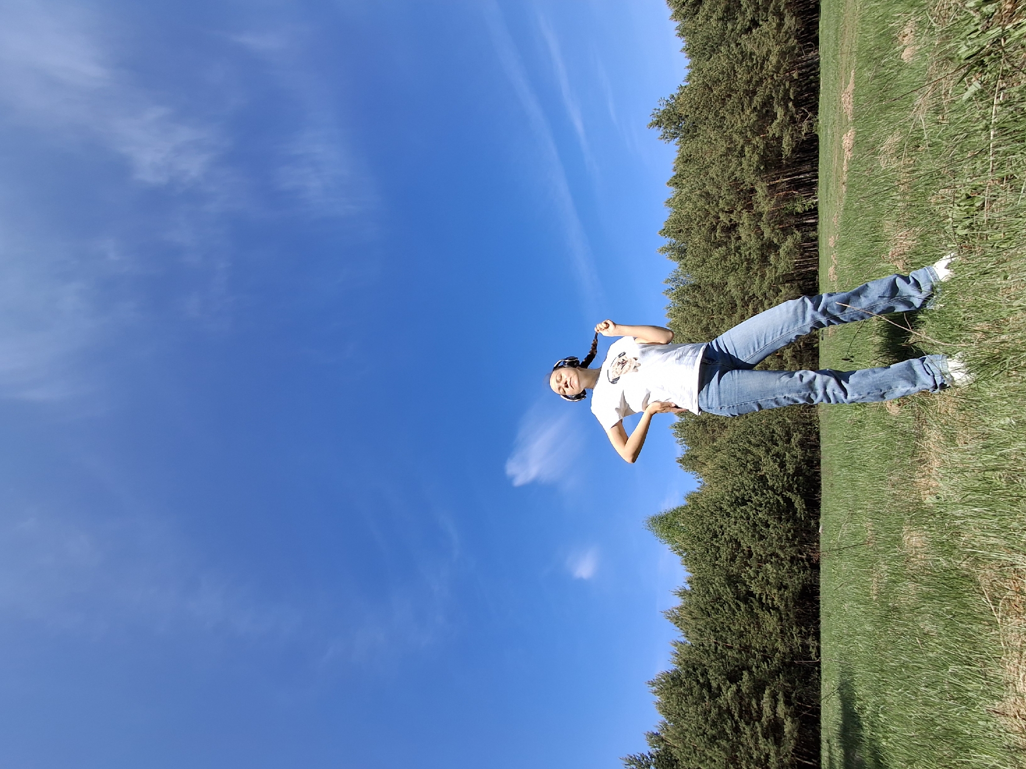 A woman is standing in a grassy field with a dense line of trees in the background. The sky is a clear, deep blue with wisps of clouds. The woman is wearing a white t-shirt, jeans, and white shoes, and has her hair in braids. She has headphones on and is holding one of her braids. The scene is captured in bright, natural sunlight. The location is the city of Chaikovsky, Russia.