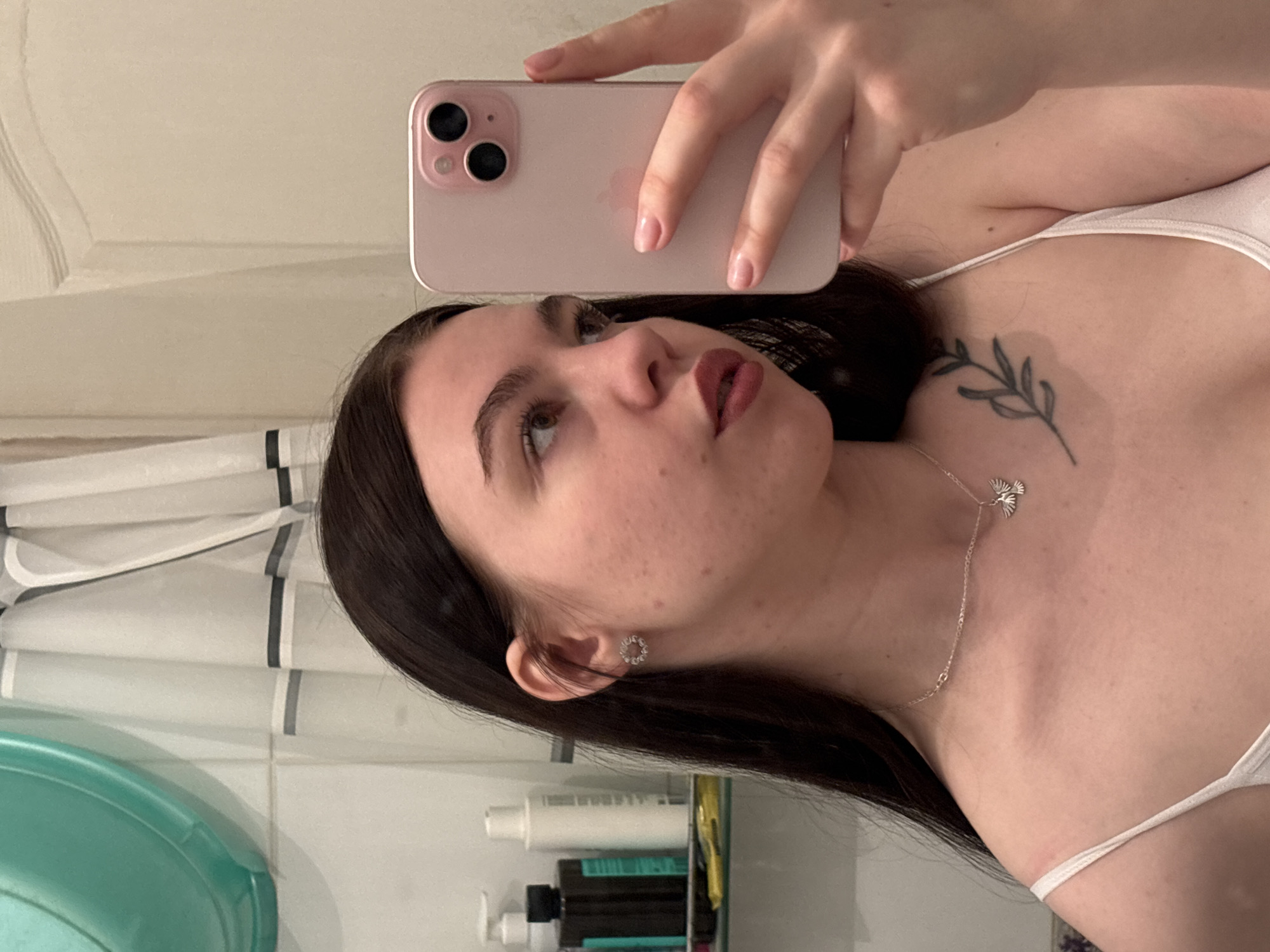 A woman with dark hair is taking a selfie in what appears to be a bathroom. She is holding a pink iPhone up to her face. She has a leaf tattoo on her upper chest and is wearing a silver necklace with a bird pendant and circular stud earrings. A partial view of a shower curtain and bathroom toiletries on a shelf are visible in the background. The image was likely taken in Perm, Russia.