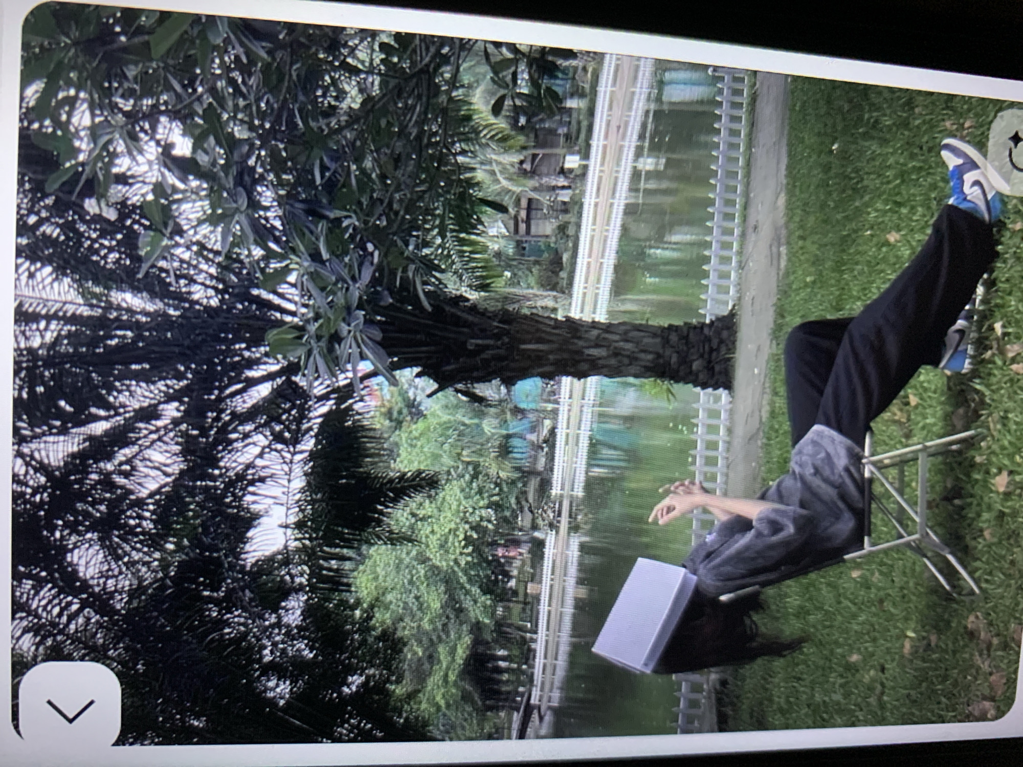 A person is seated on a metal folding chair outdoors, with a book placed on their head. They are wearing a gray t-shirt and dark pants, with blue and white sneakers. The person is positioned on a grassy area in front of a body of water, possibly a pond or lake, with a white picket fence bordering the water. In the background, there are trees and some structures. The location is Lymanka, Ukraine.
